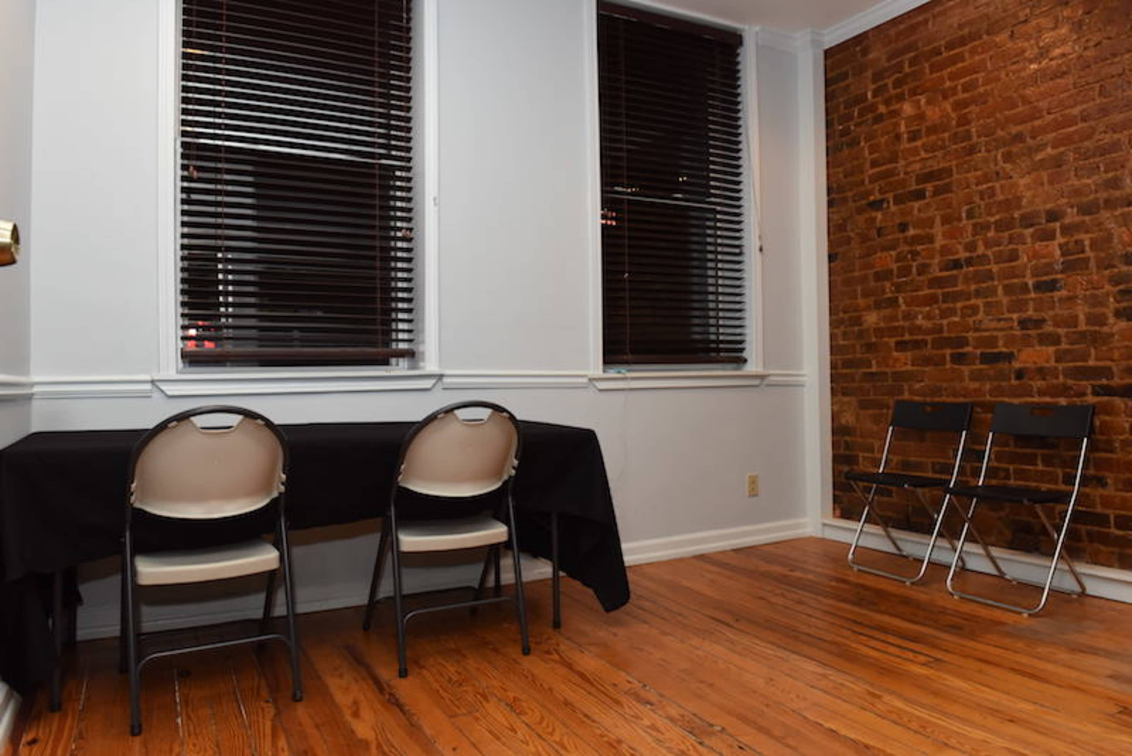 The room features two empty chairs at a table beside a brick wall and three windows with blinds.