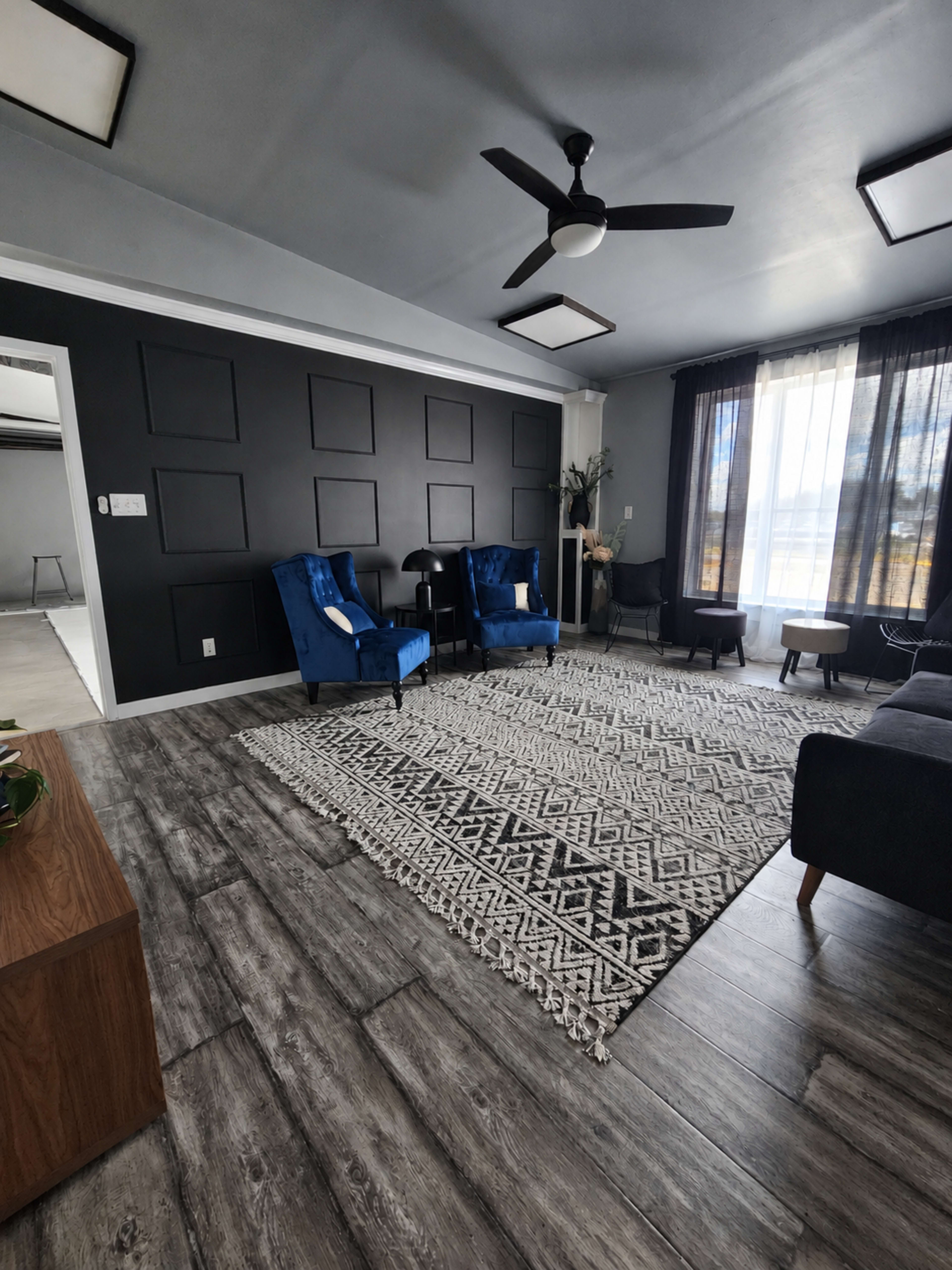 The image shows a modern living room with two blue velvet chairs, a patterned rug, and dark walls featuring geometric designs.