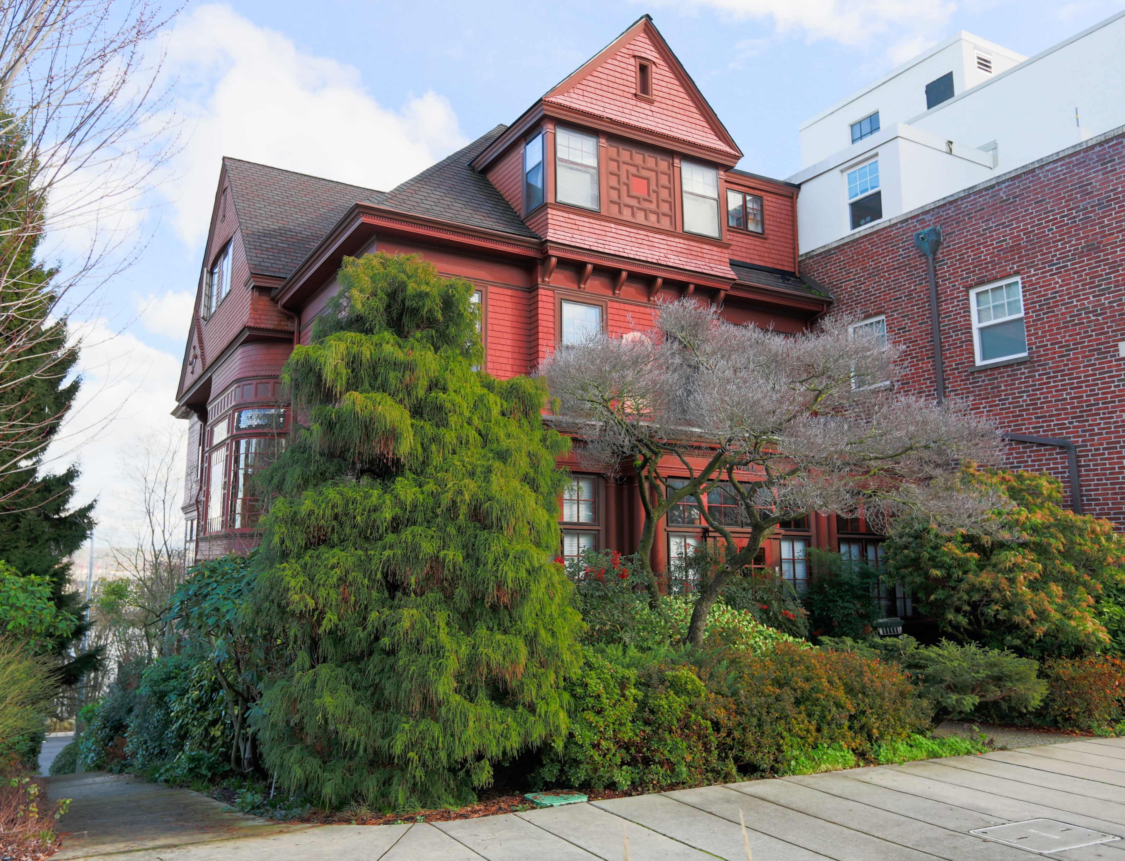 A red Victorian-style house with intricate architectural details is surrounded by lush greenery and trees.
