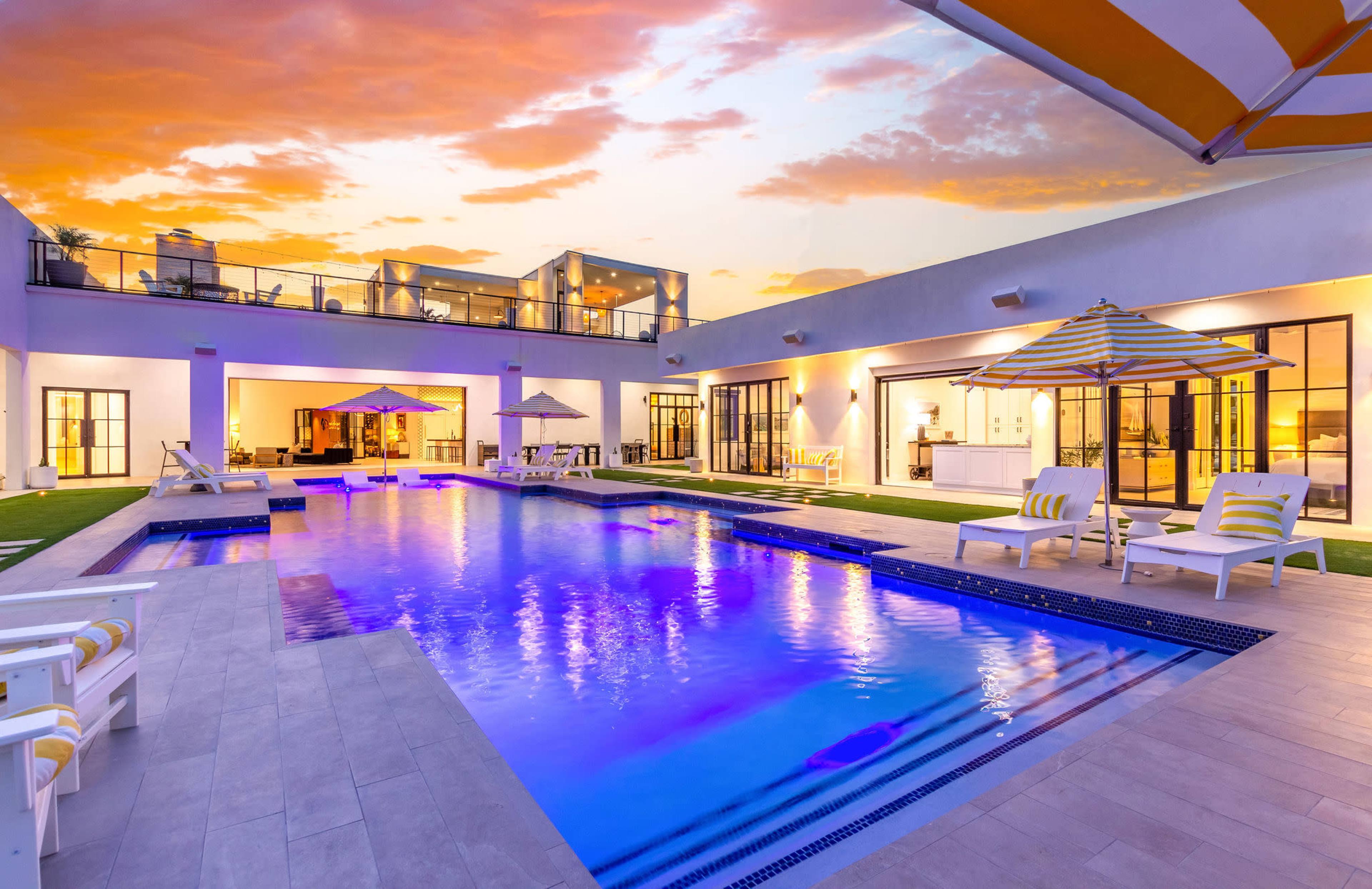 A modern outdoor pool area features lounge chairs, umbrellas, and a backdrop of a sunset sky.
