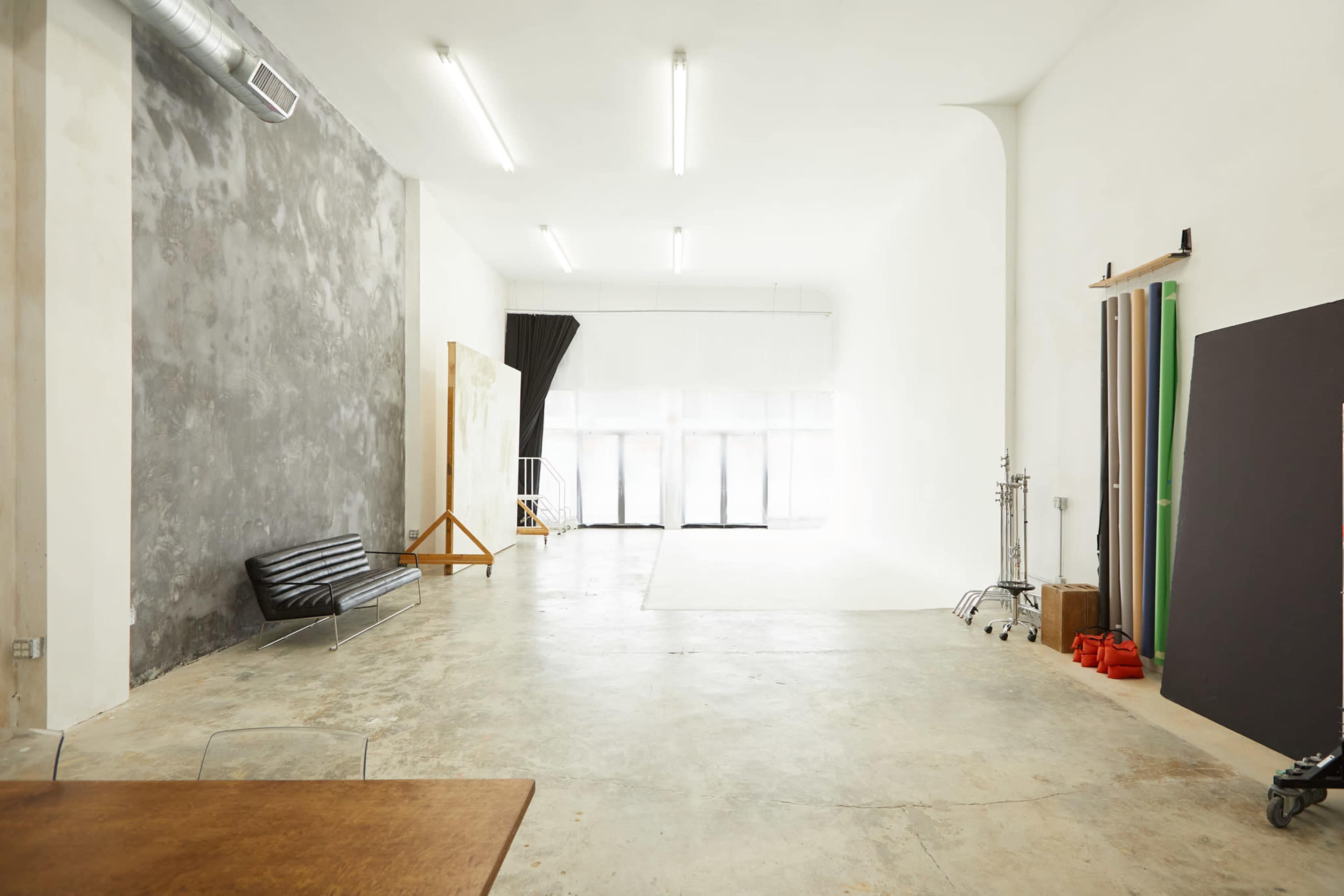 A spacious, minimalist studio with concrete floors, large windows, and a variety of colorful backdrops leaning against the wall.