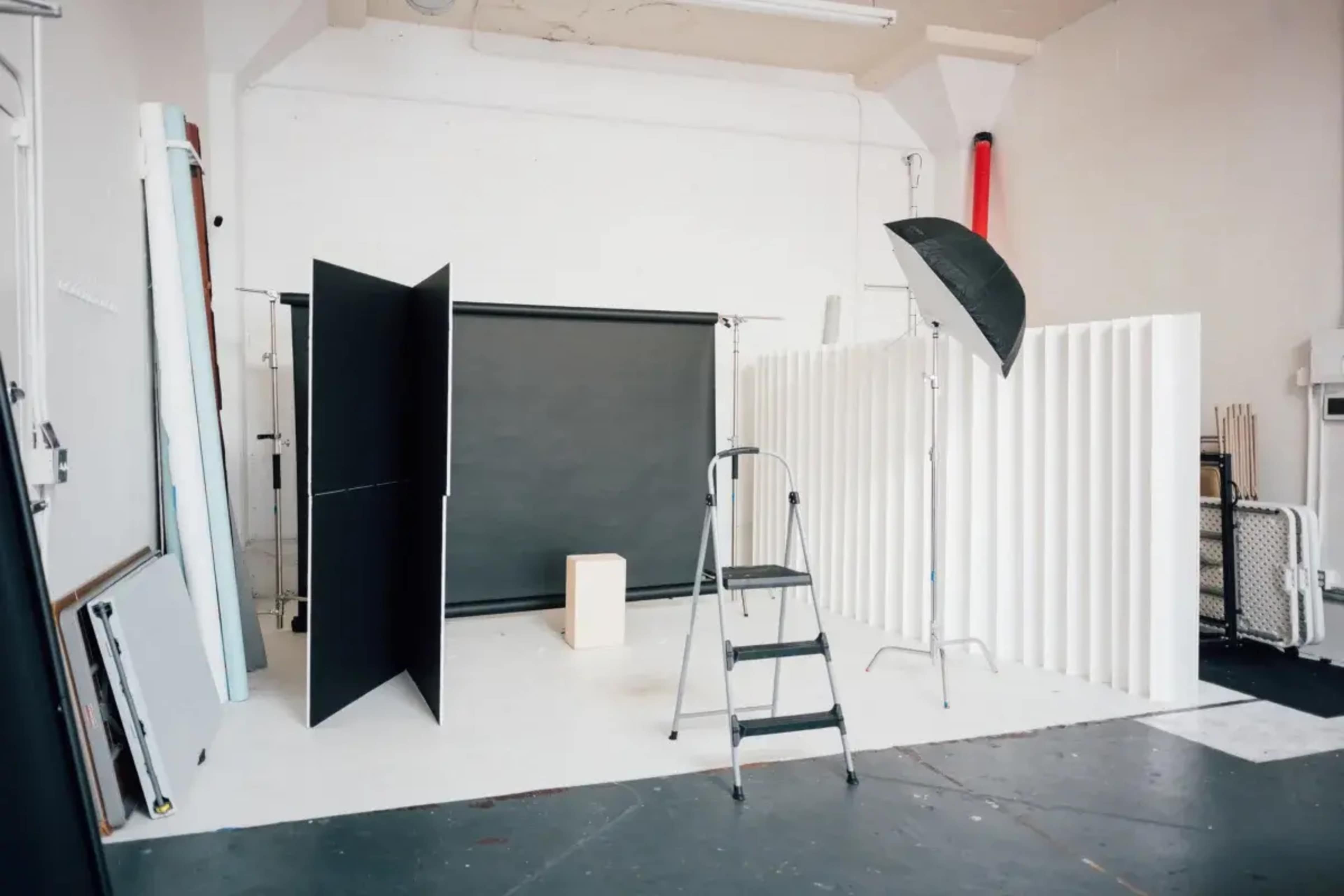 The image shows a photography studio setup with a plain backdrop, a step ladder, lighting equipment, and white and black screens arranged around a central wooden box.