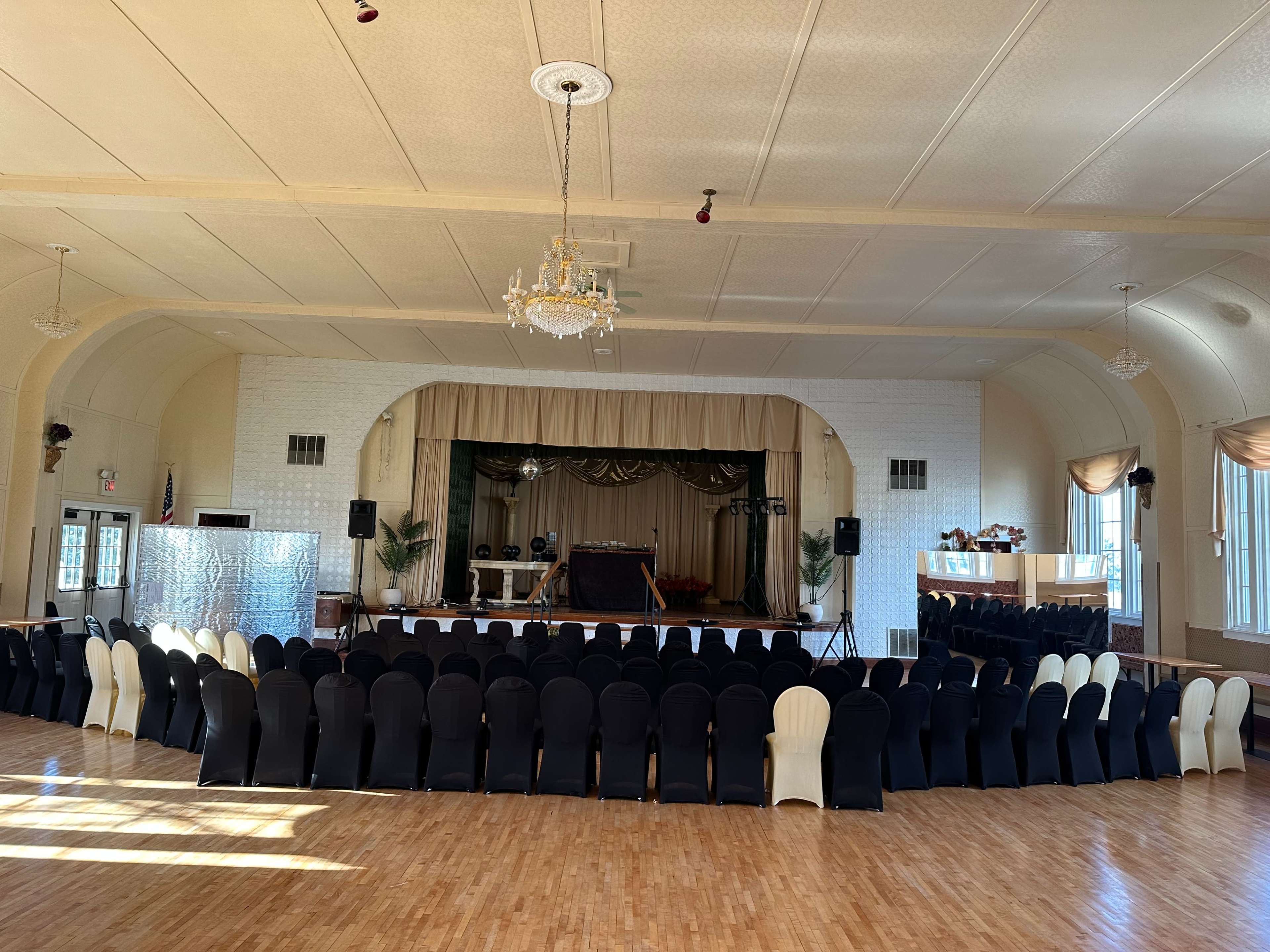 A spacious event hall set up with black and cream chairs arranged in a circular layout facing a stage with a dark curtain and sound equipment.