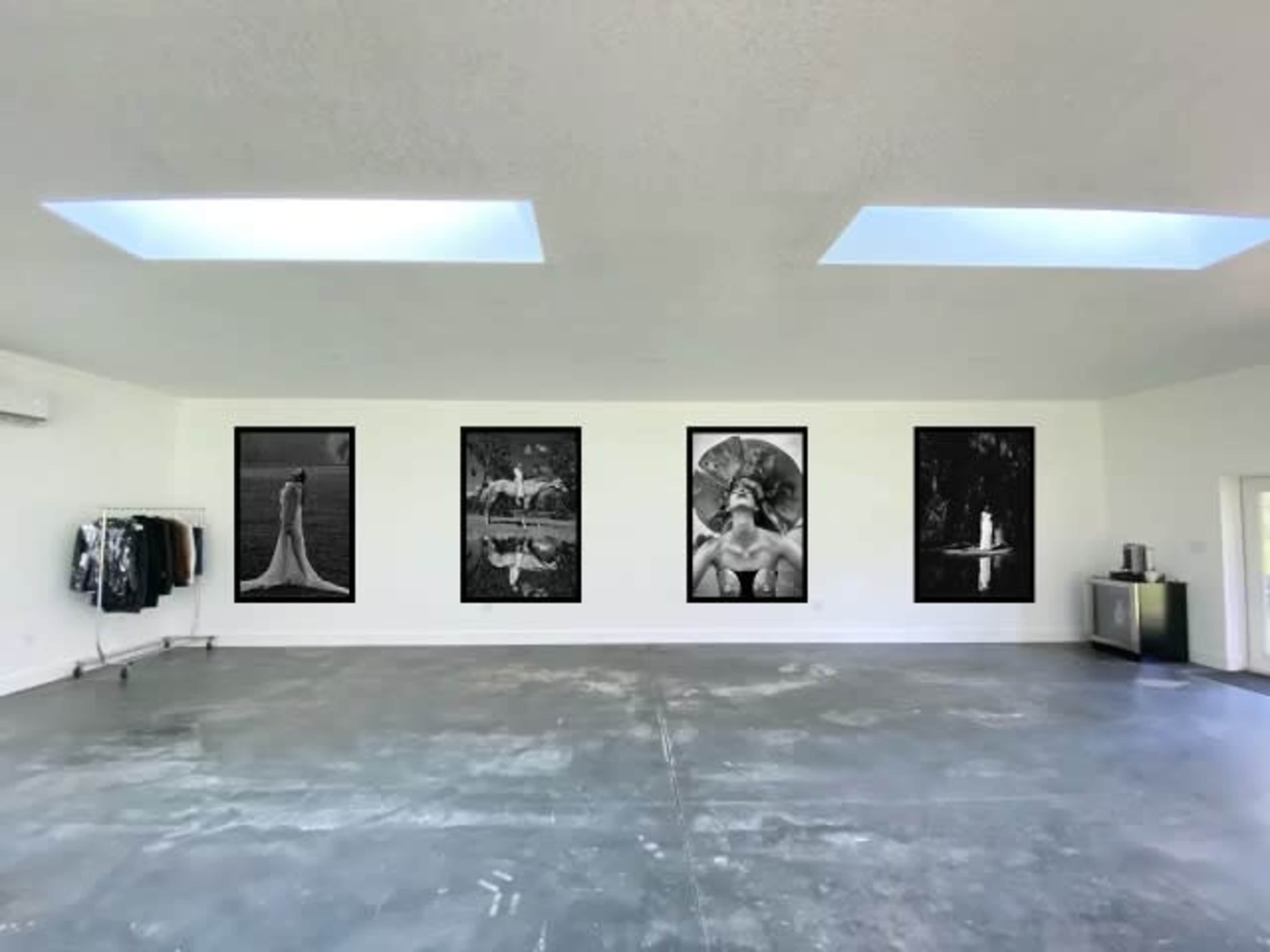 A spacious, minimalist art gallery featuring four black-and-white photographs on the walls, with clothing displayed on a rack in the corner.