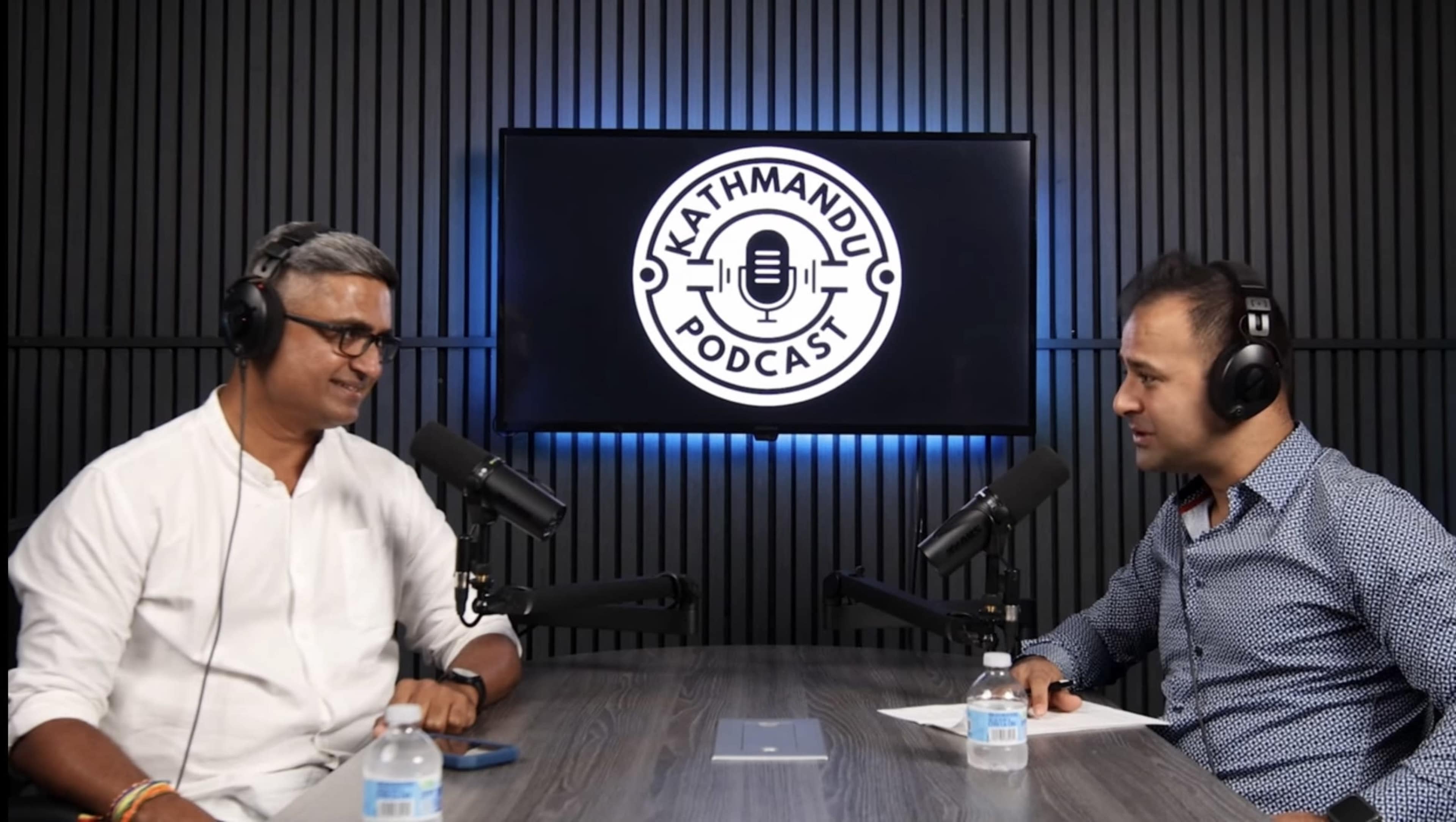 Two men are seated at a table with microphones in front of them, discussing in a podcast studio featuring a backlit logo that reads "Kathmandu Podcast."