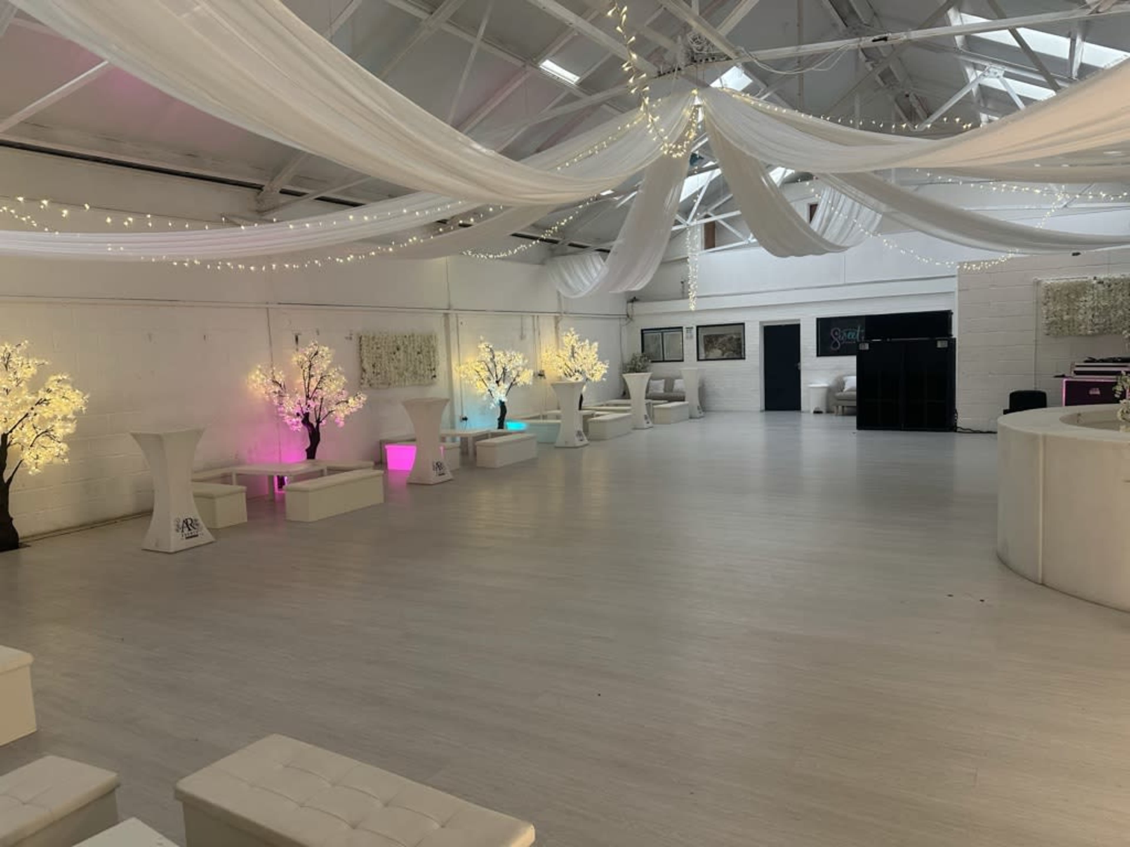 The image shows a spacious indoor venue with white walls, a light-colored floor, and decorative illuminated trees.