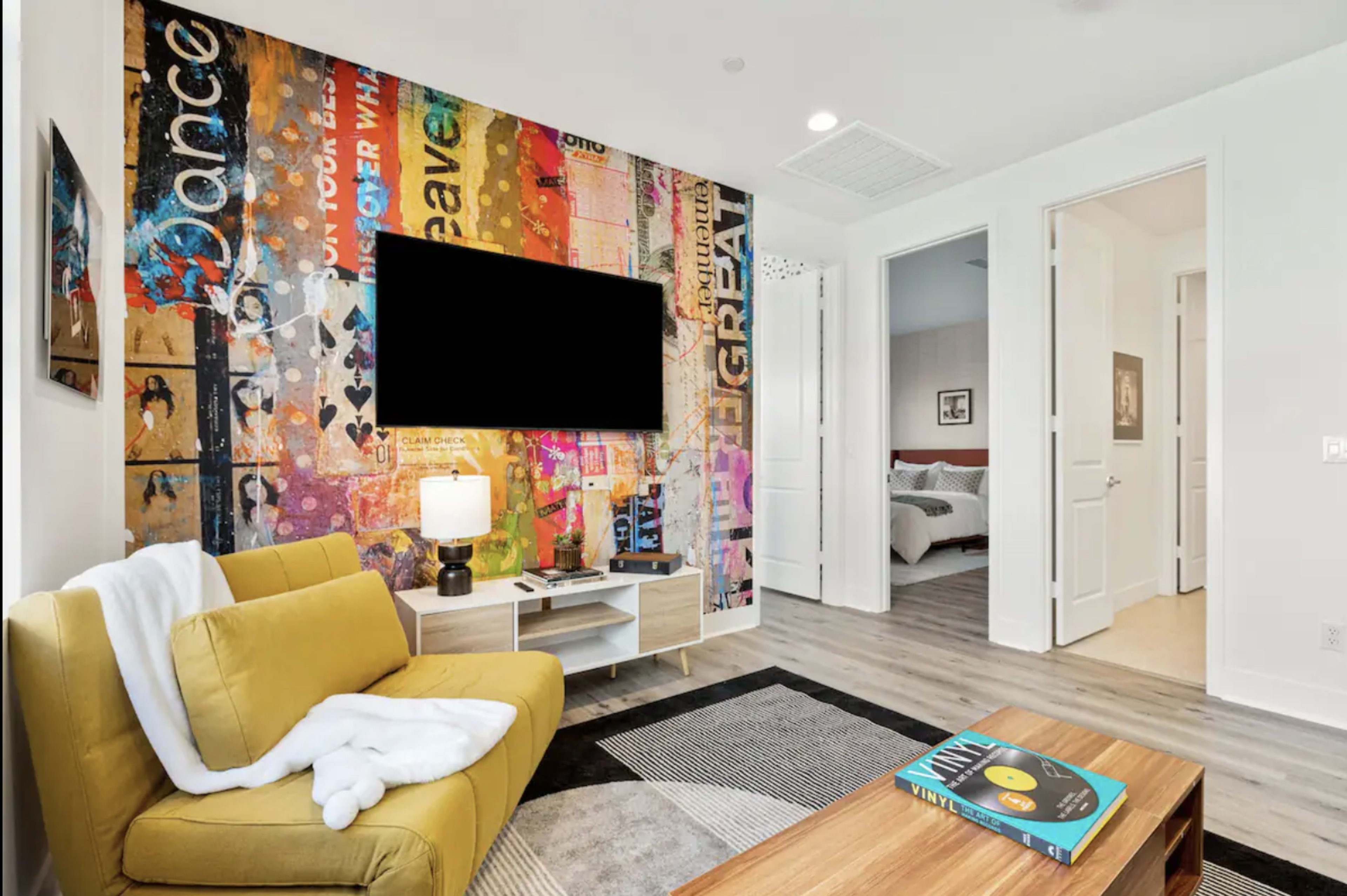 A living room features a yellow sofa, a coffee table with a vinyl record, and a wall mural with colorful graphics, alongside a mounted television.