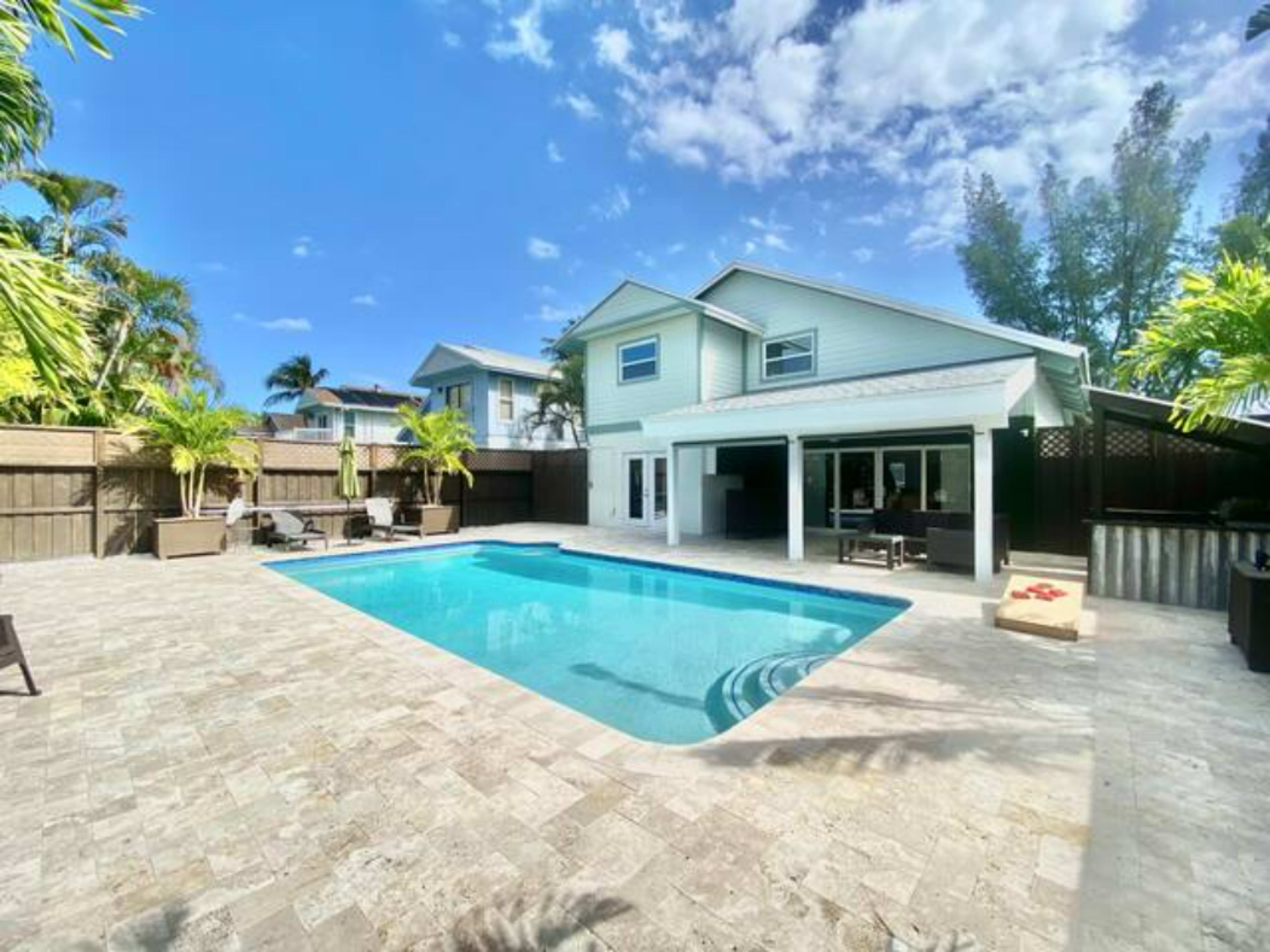 A backyard with a swimming pool, surrounded by stone pavement and tropical landscaping, with a two-story house in the background.