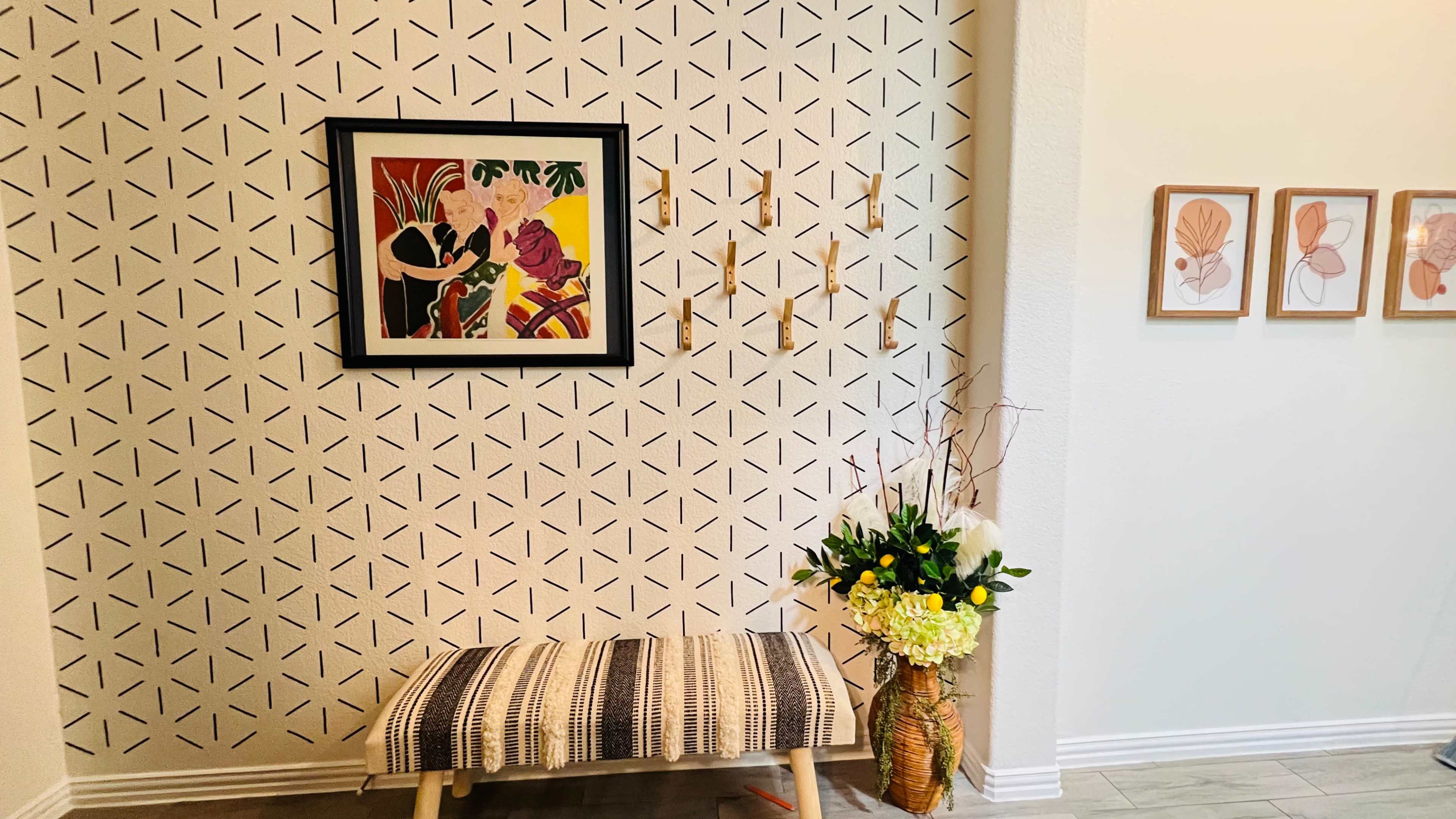 A modern entryway featuring a patterned accent wall with a framed art piece, a wooden bench, and a decorative flower arrangement.