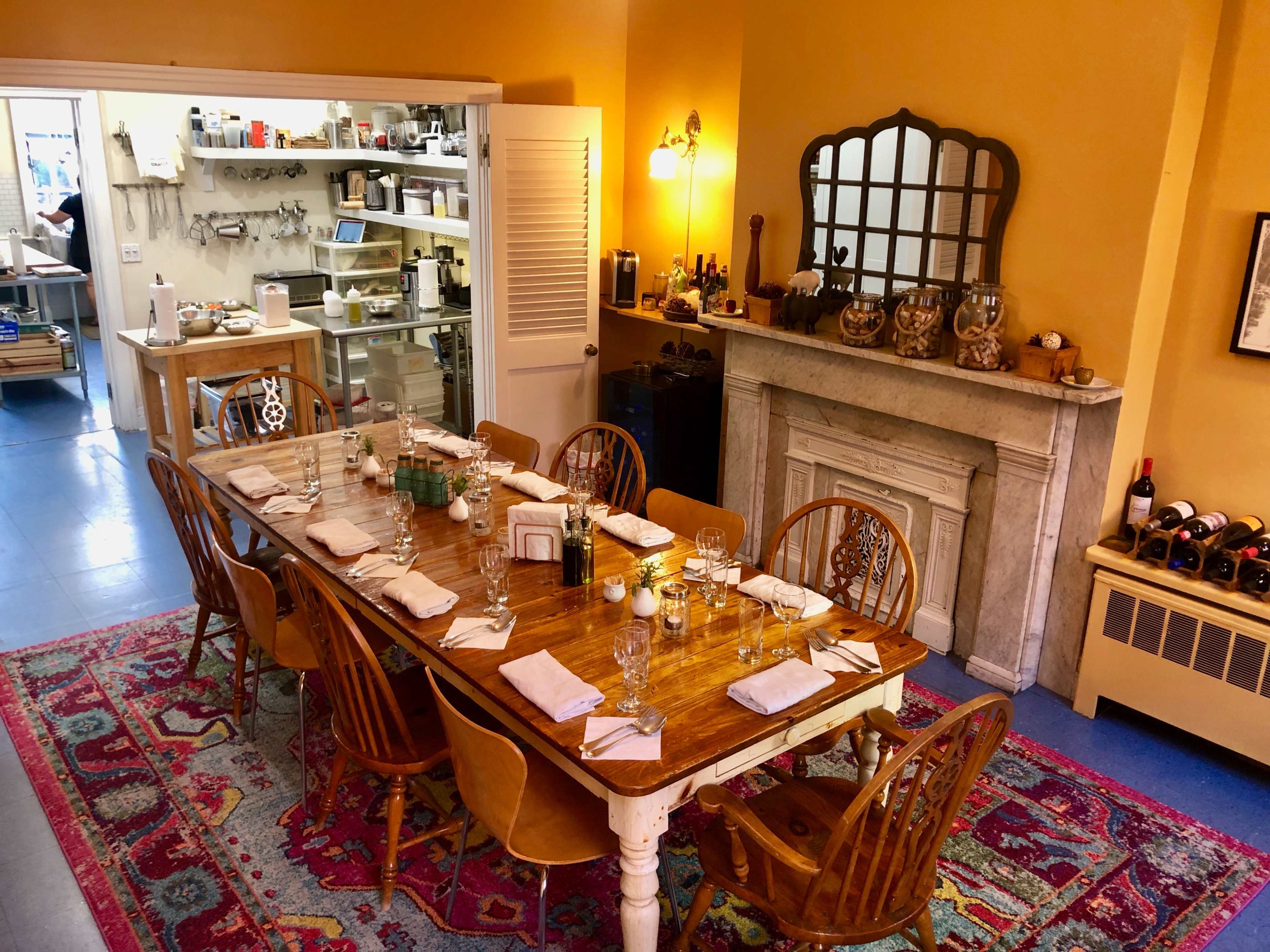 A long wooden dining table is set with plates and glasses in a warmly lit dining room, with a kitchen area visible in the background.
