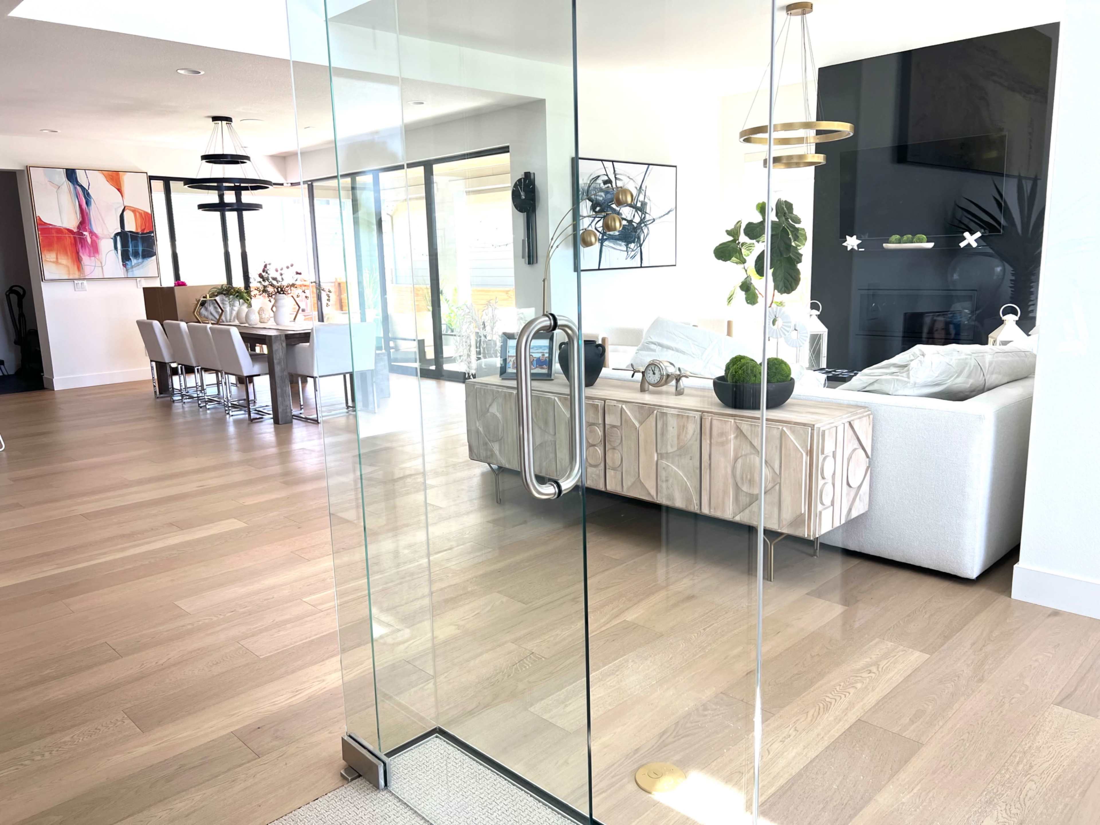 The image shows a modern, open-concept living space with a glass door leading to a lounge area featuring a light-colored sofa and a dining area in the background.