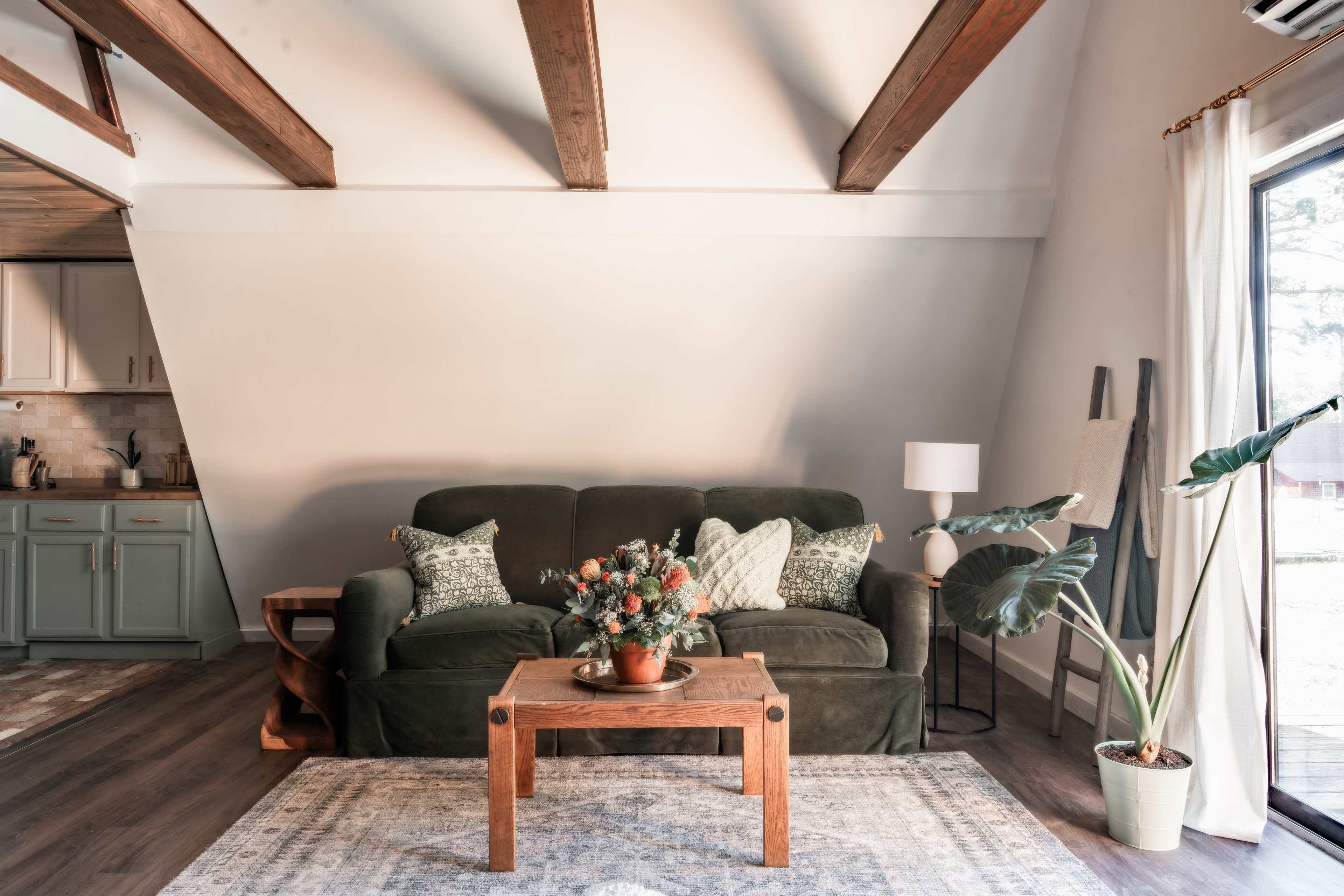 A green sofa with patterned cushions sits in a brightly lit room featuring wooden beams, a coffee table with flowers, and a window with a potted plant nearby.