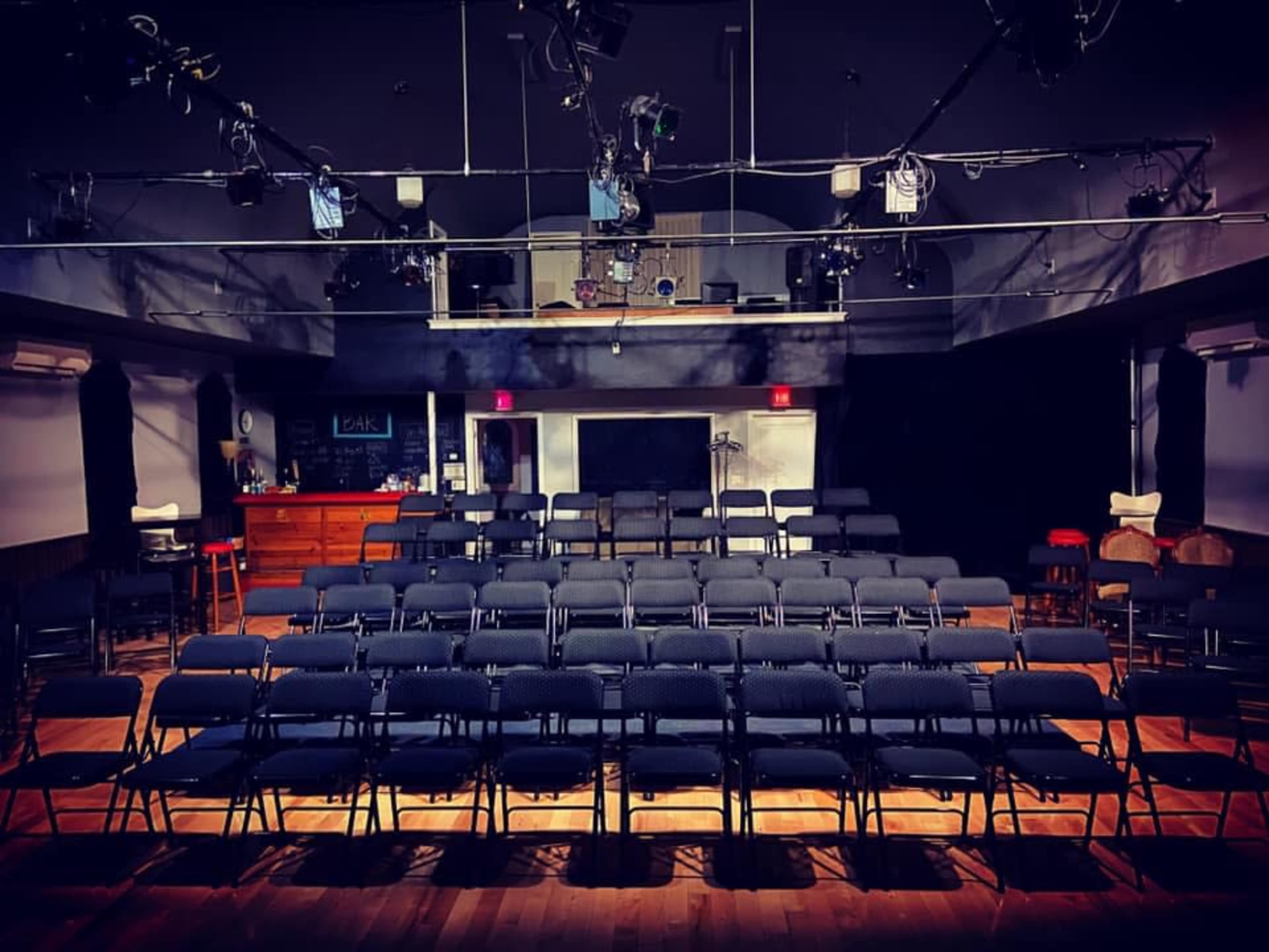 A theater setup with rows of black folding chairs facing a stage, illuminated by overhead stage lights.
