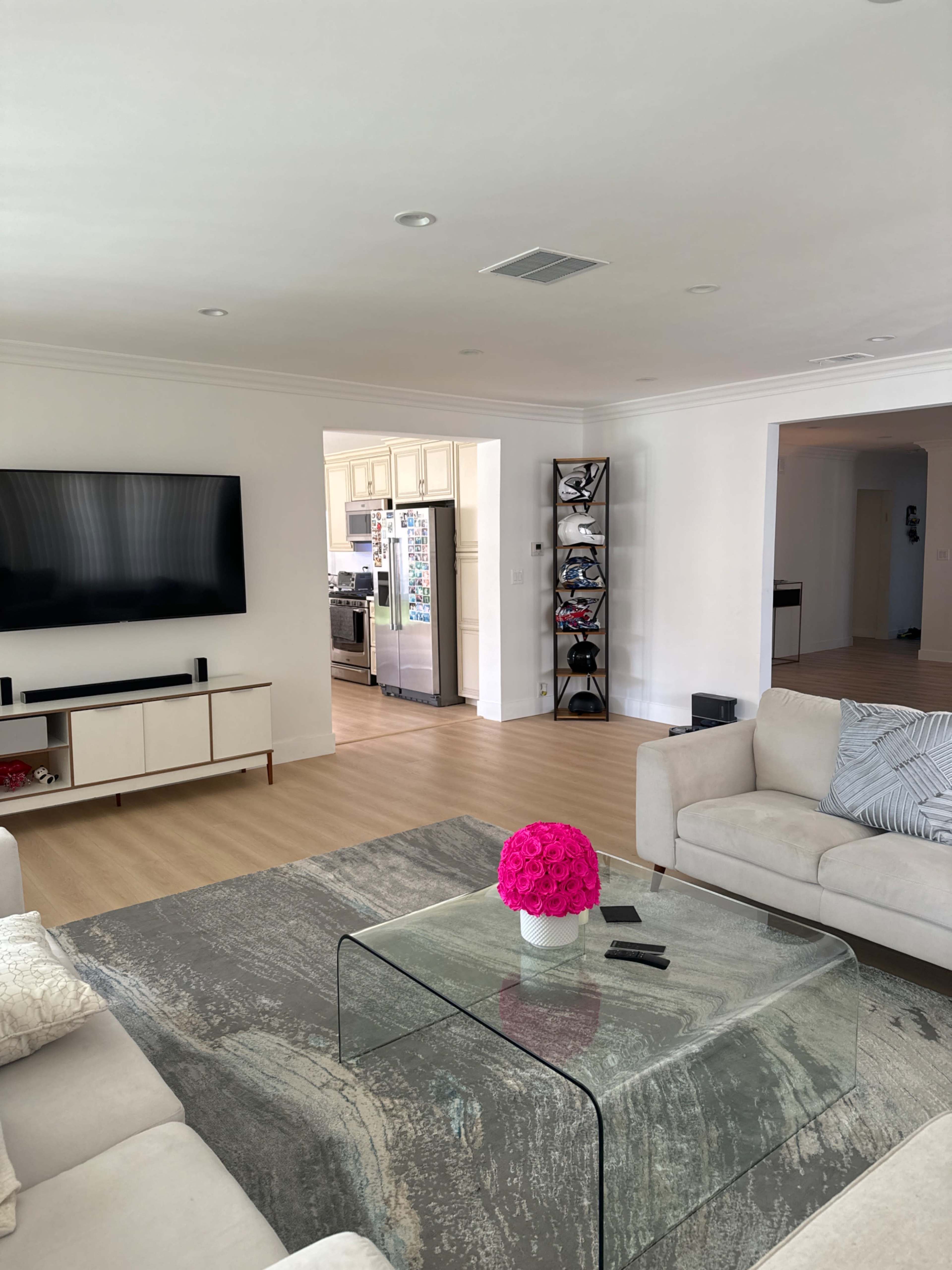 The living room features a large TV mounted on the wall, a glass coffee table with a pink flower arrangement, and a view of an adjoining kitchen.