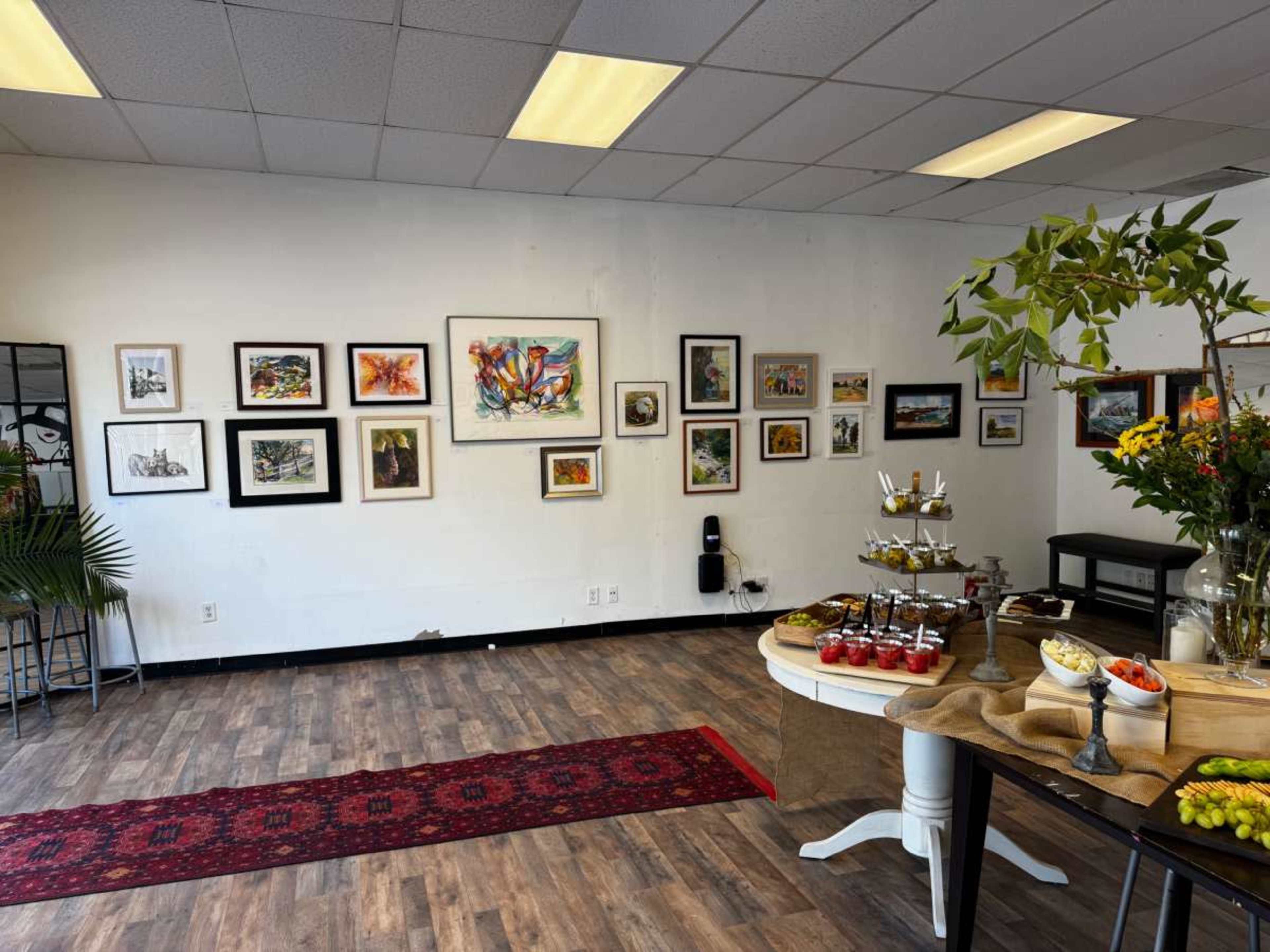 The image shows an art gallery interior with framed artwork on the walls, a table adorned with fruit and pastries, and a decorative rug on the wooden floor.