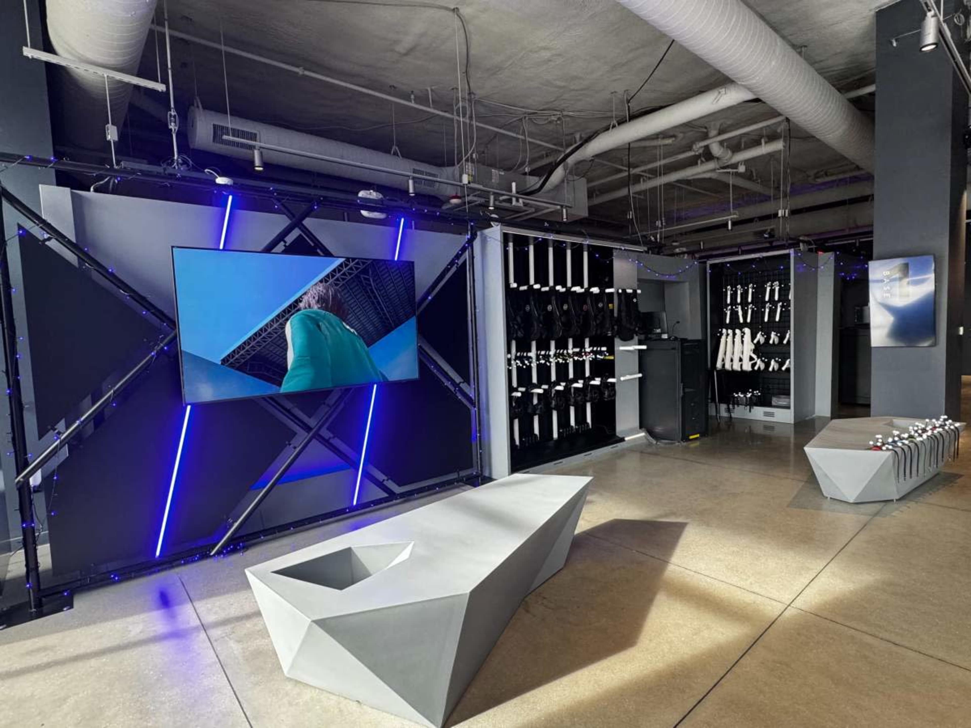 The image shows a modern interior space featuring a large screen displaying a video, with sleek geometric furniture and black storage units in the background.