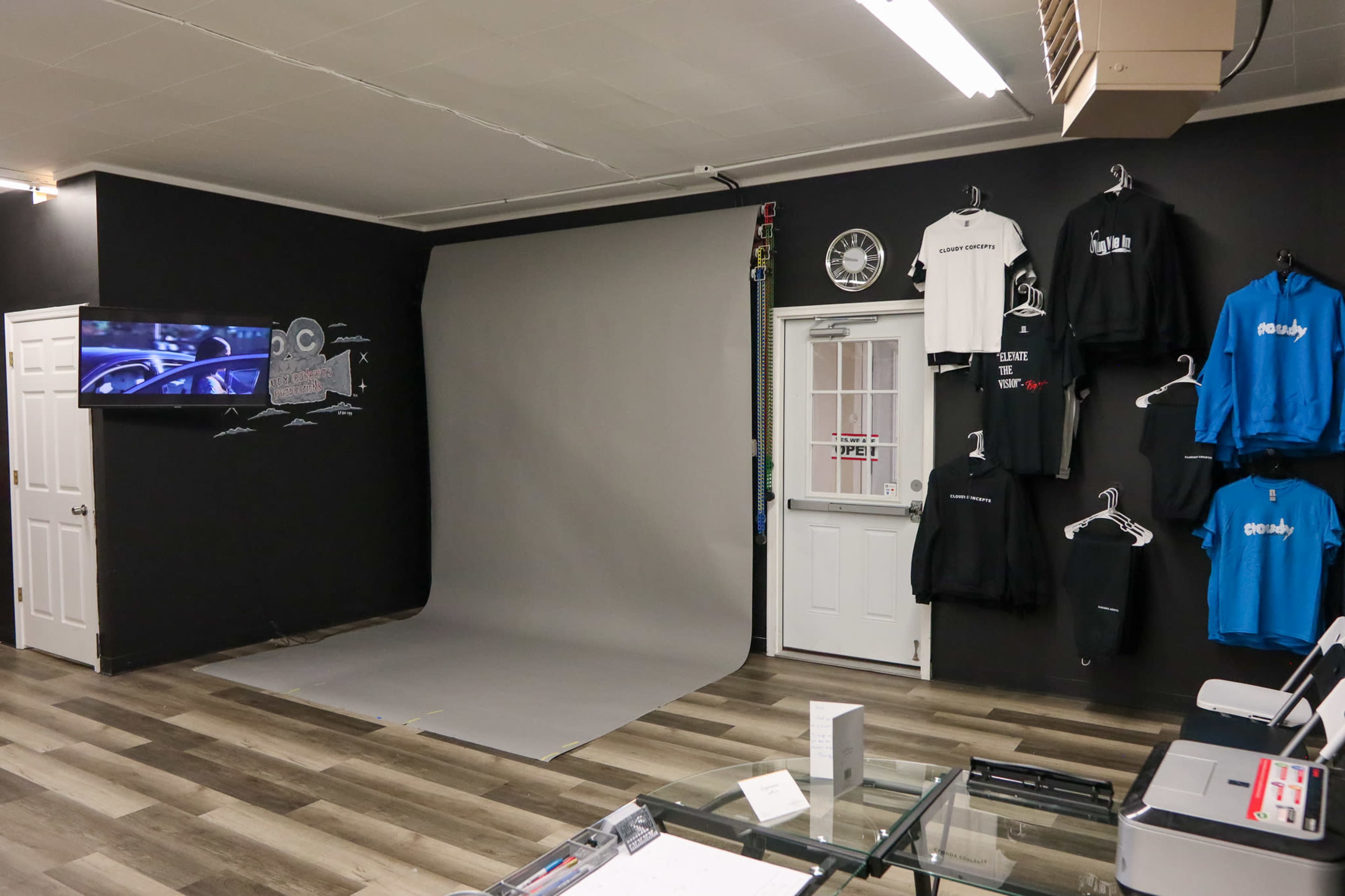 An interior space with a gray backdrop for photography, clothing displayed on hangers, and a television mounted on the wall.