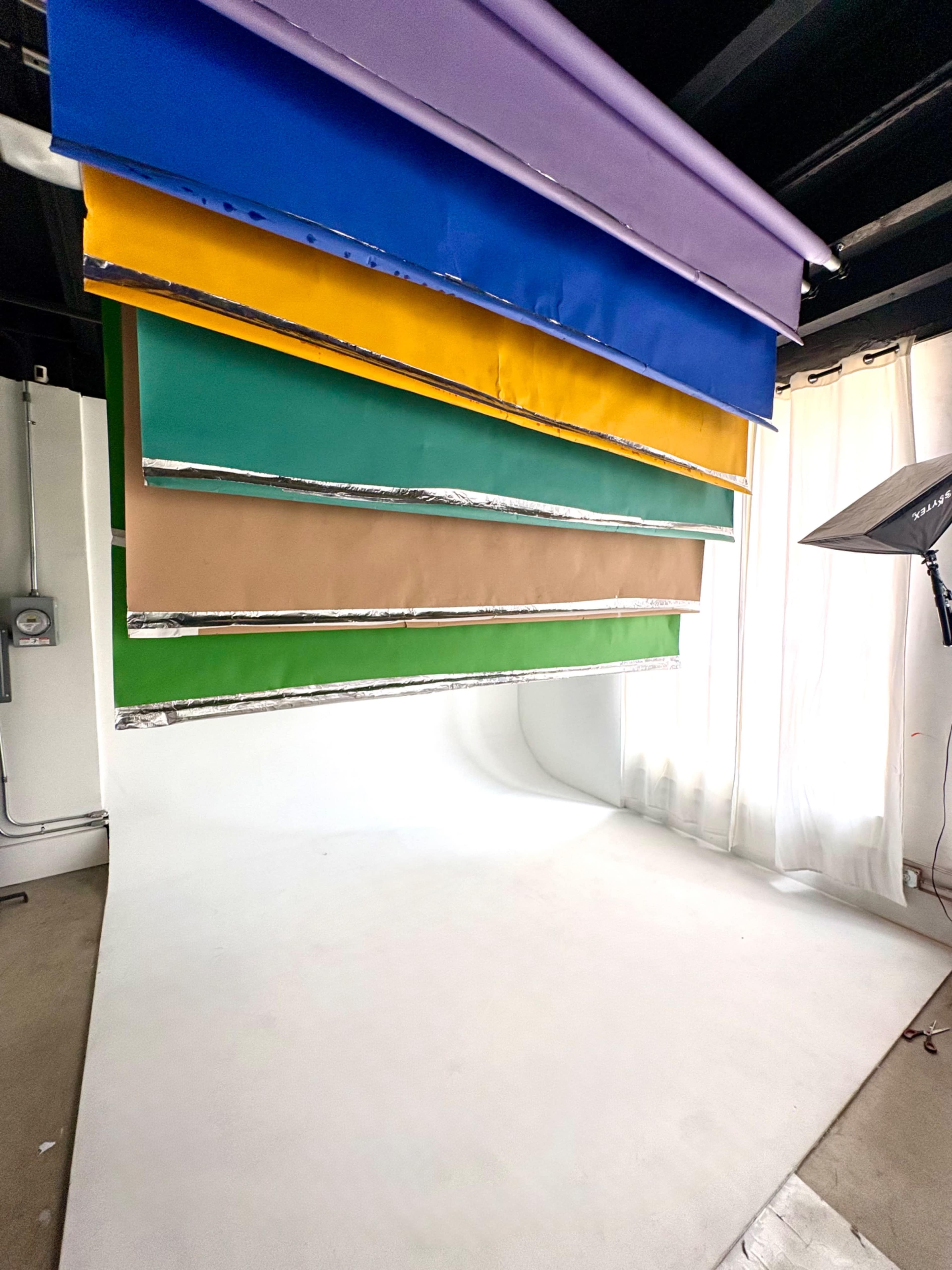 The image shows a studio setting with several color backdrops hanging vertically and a white backdrop on the floor, illuminated by a soft light source.