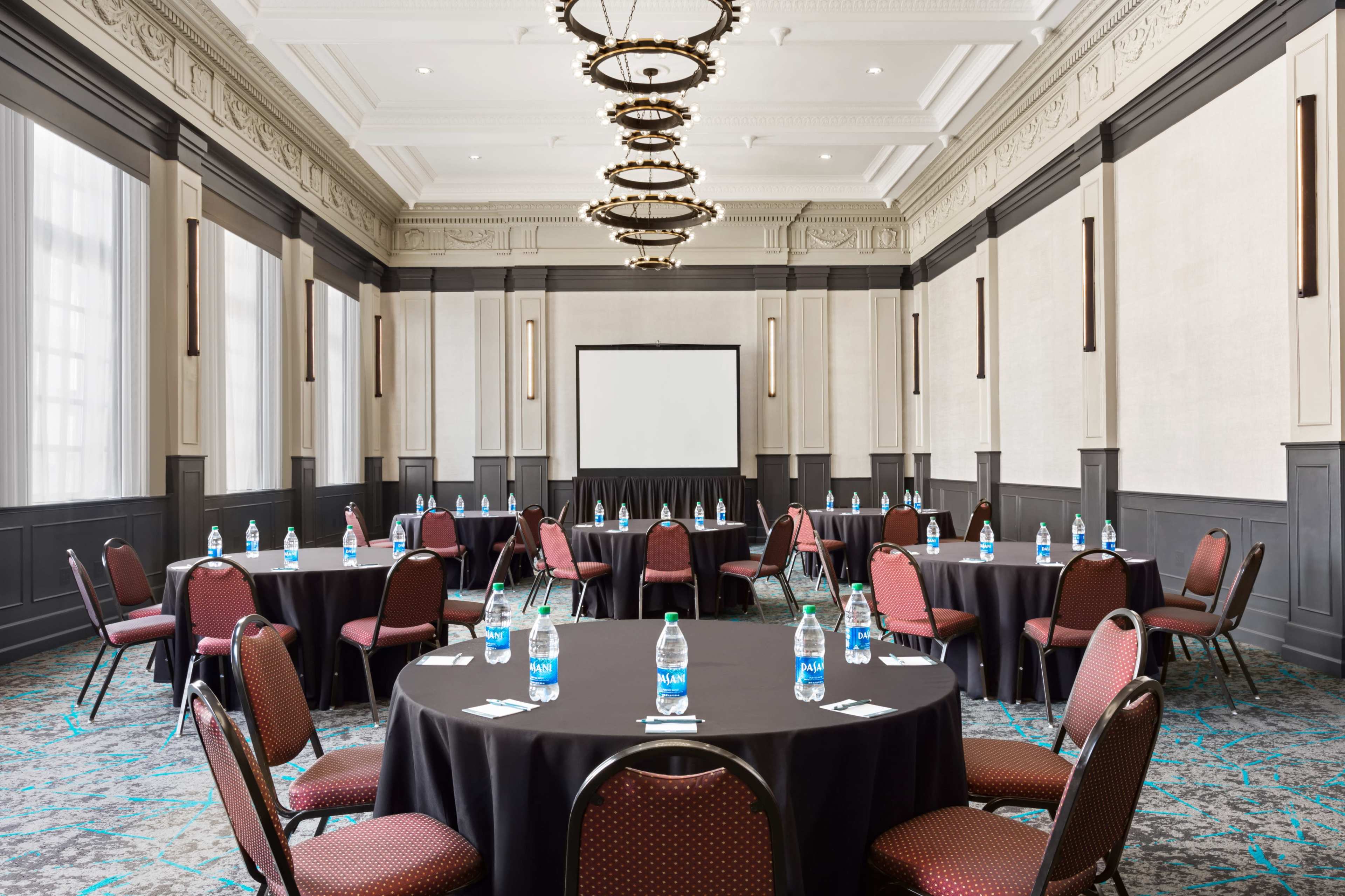 The image shows a conference room set up with several round tables, each surrounded by chairs, and featuring water bottles on the tables, with a projector screen at the front.
