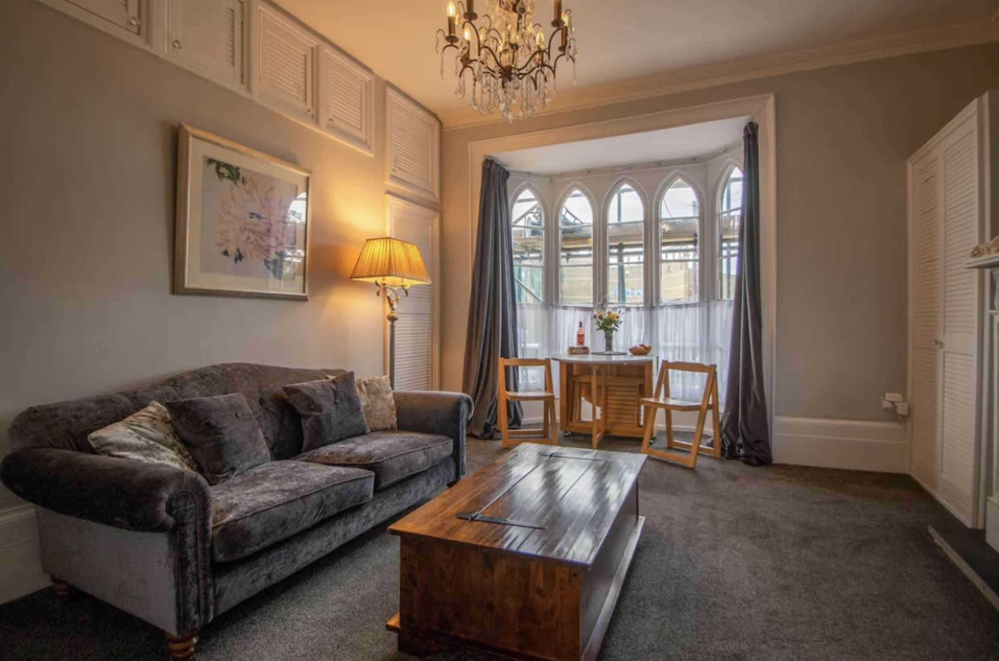 The image shows a cozy living room with a gray sofa, a wooden coffee table, and a dining area featuring two chairs and a small table next to large arched windows.