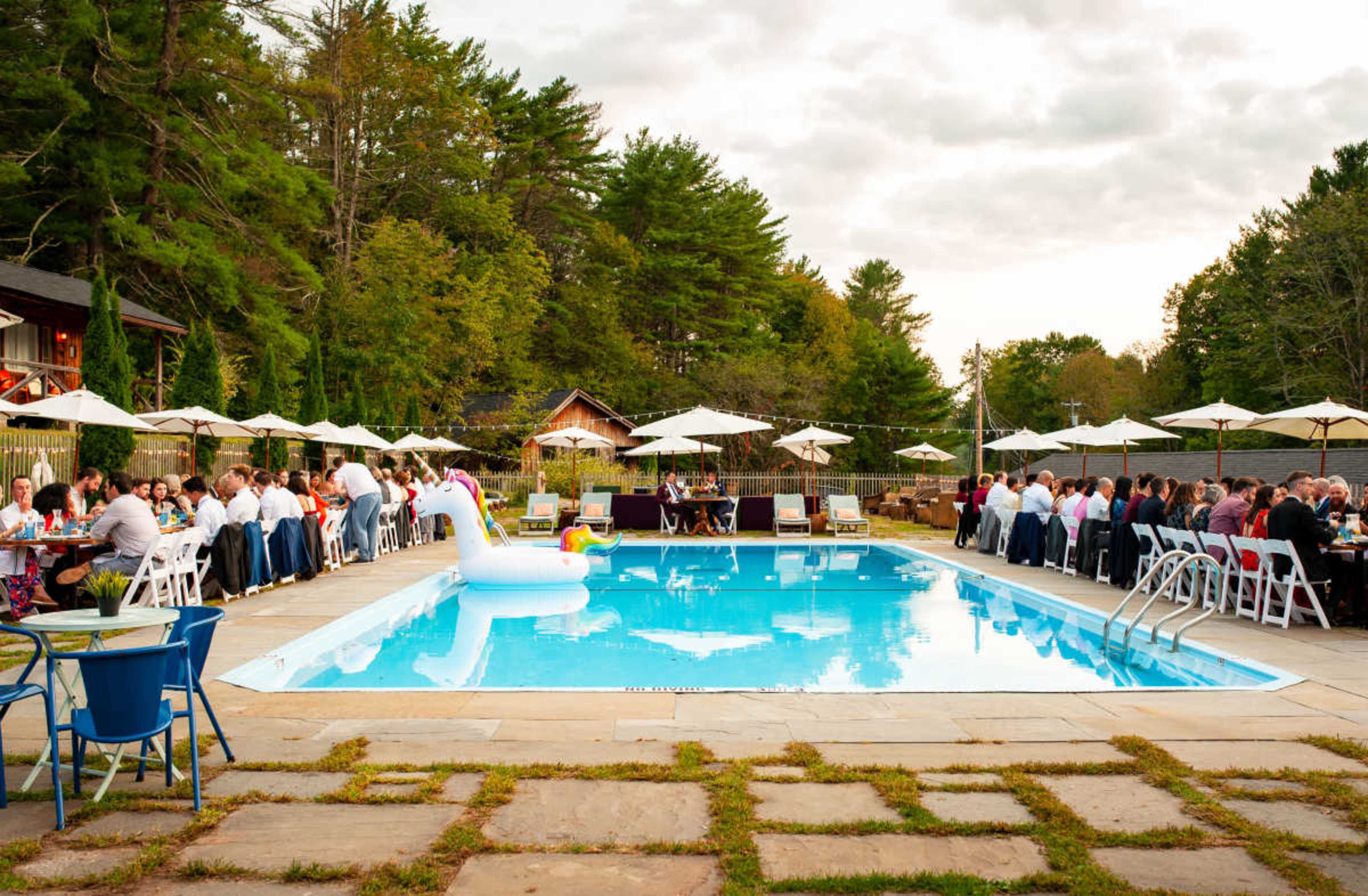 A large outdoor gathering takes place by a swimming pool, with tables set up for guests and a white swan-shaped float in the water.