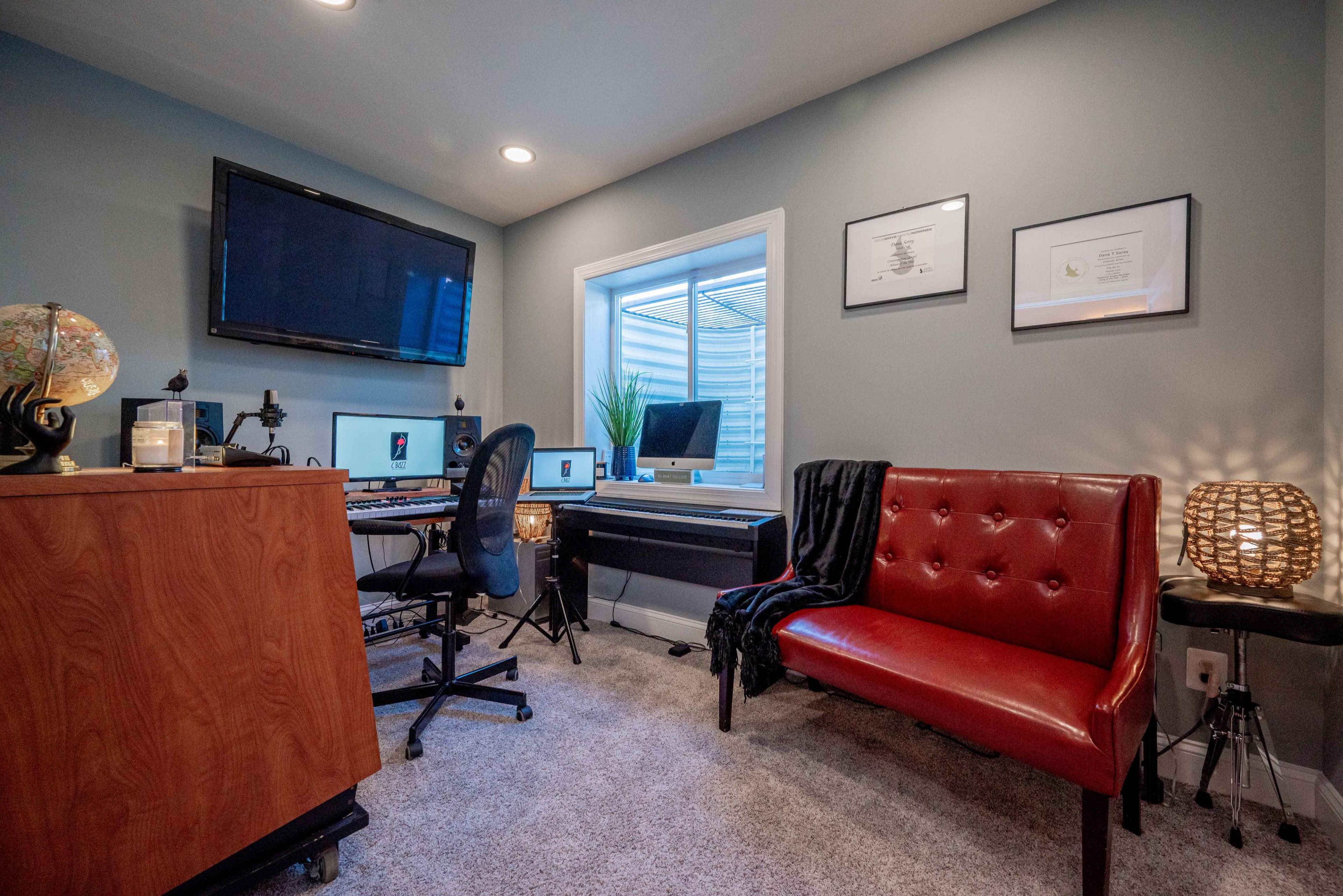 A small home office with a desk, computer, keyboard, a red couch, and a window revealing a plant outside.