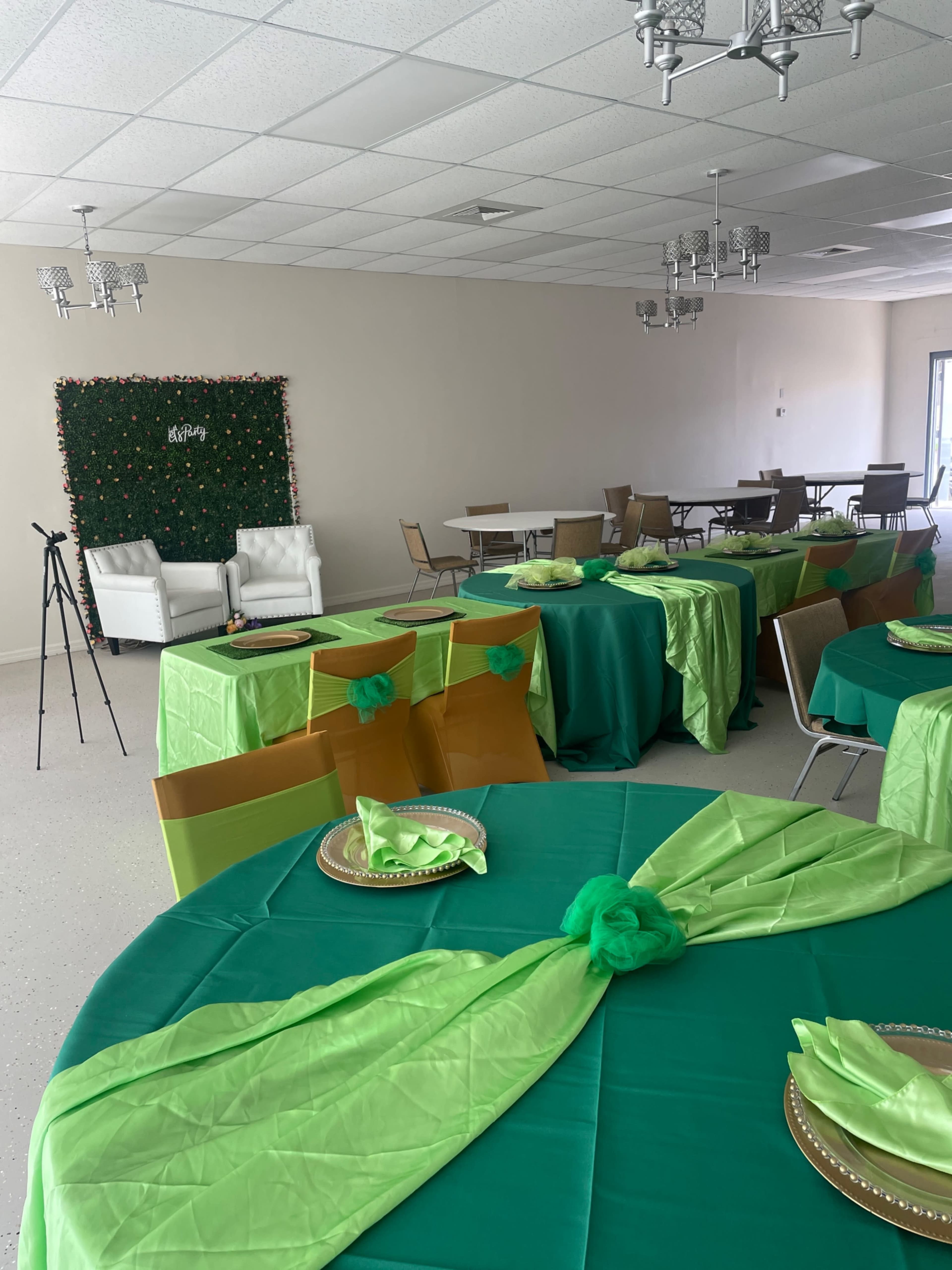 A spacious event room with green and gold tablecloths draped over several tables, neatly set with plates and napkins, and featuring white chairs in the background.