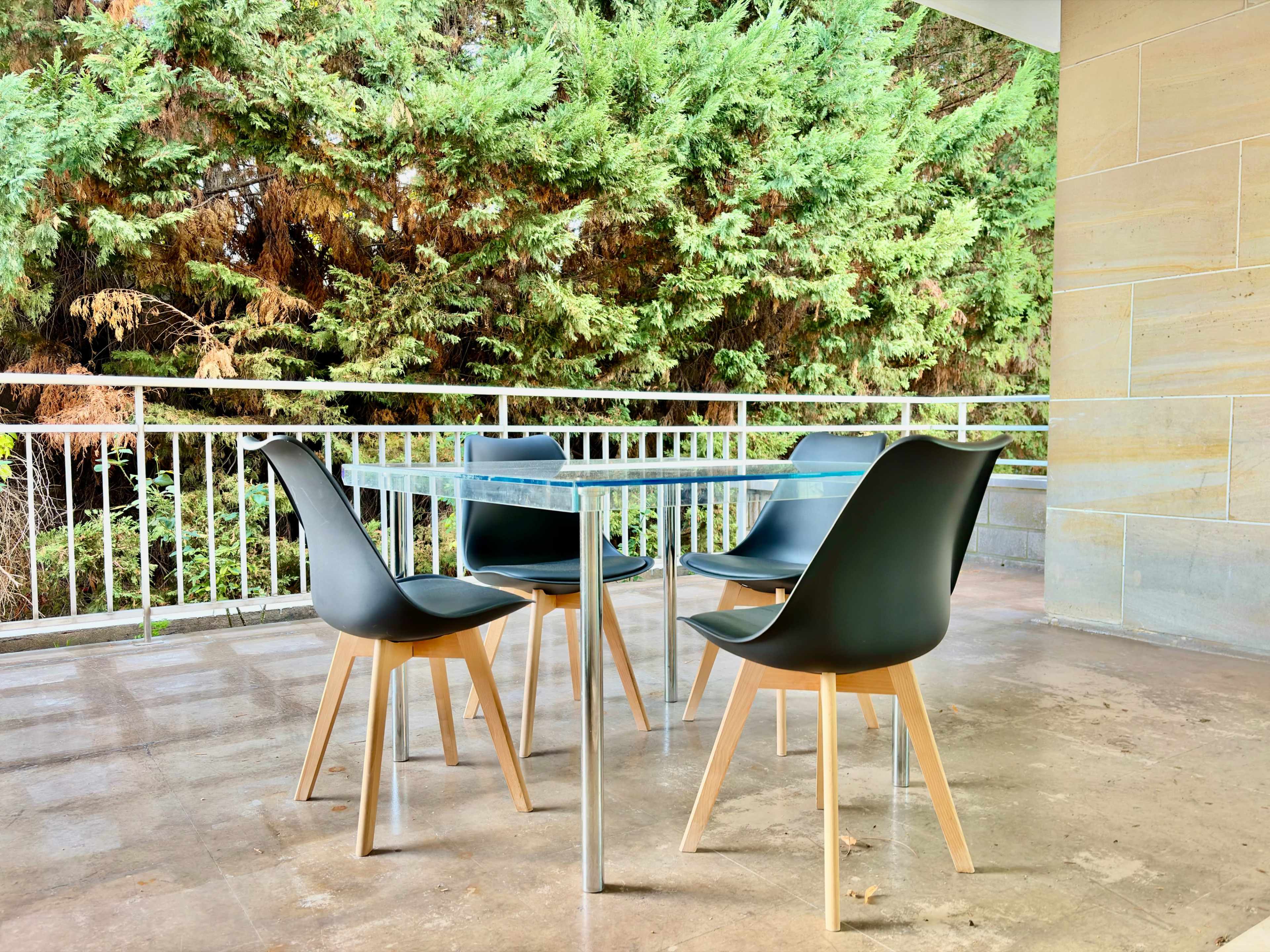 A glass table with four black chairs is positioned on a patio overlooking a backdrop of dense green foliage.