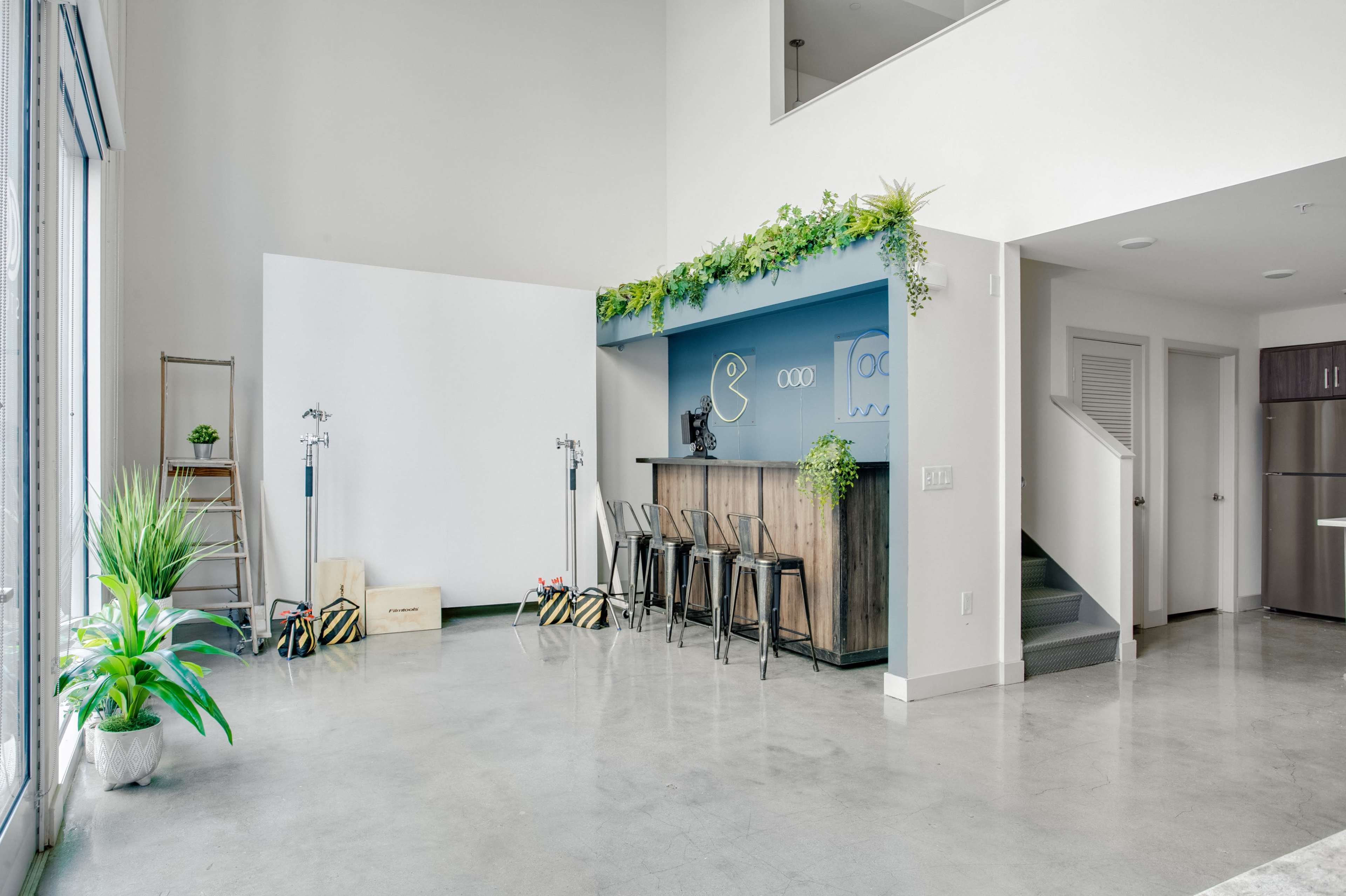 The interior features a modern open space with a bar area, black stools, and greenery on the wall, complemented by concrete flooring and large windows.