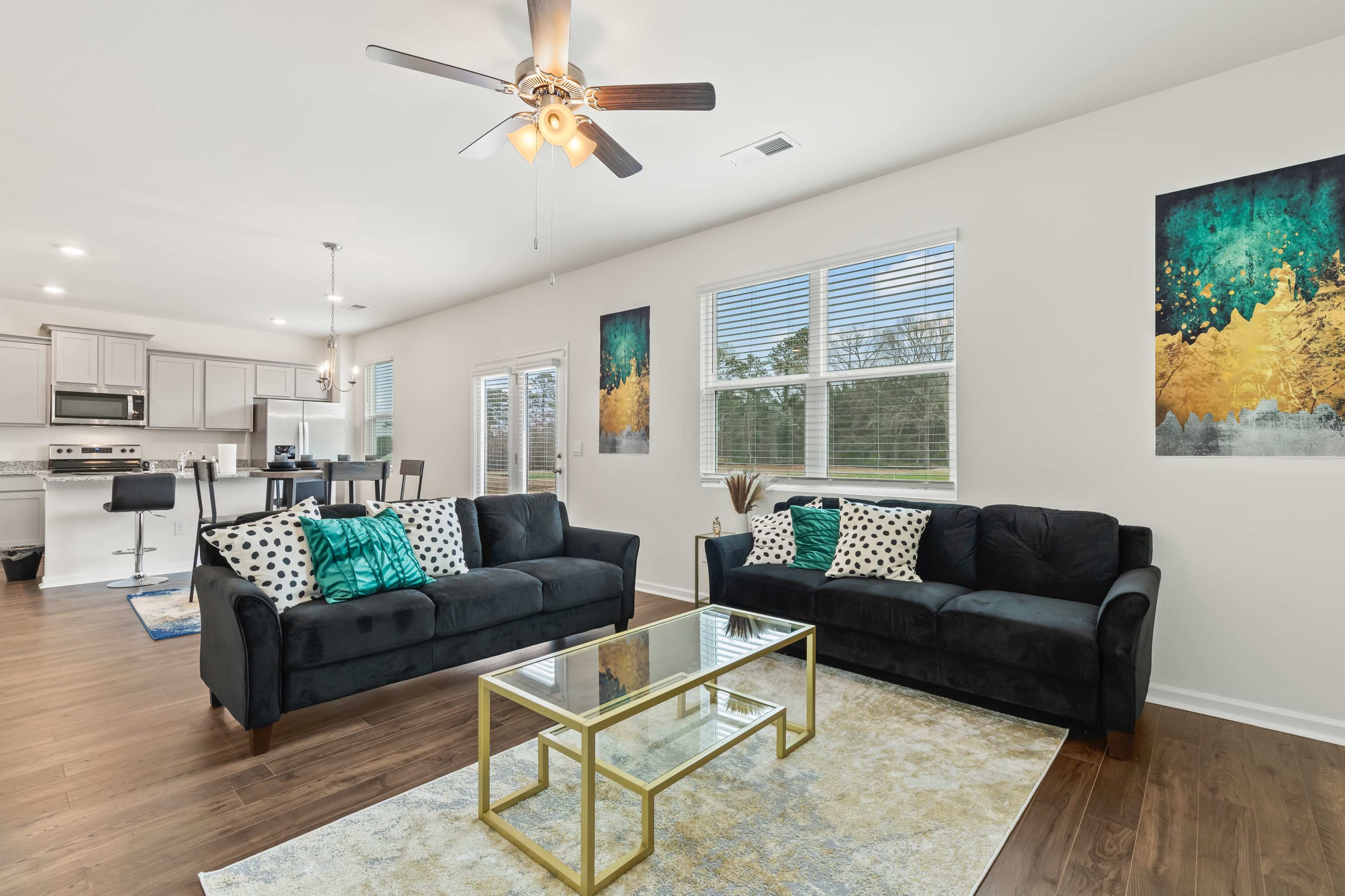 A modern living room with two dark sofas, a glass coffee table, and decorative artwork on the walls, illuminated by natural light from large windows.