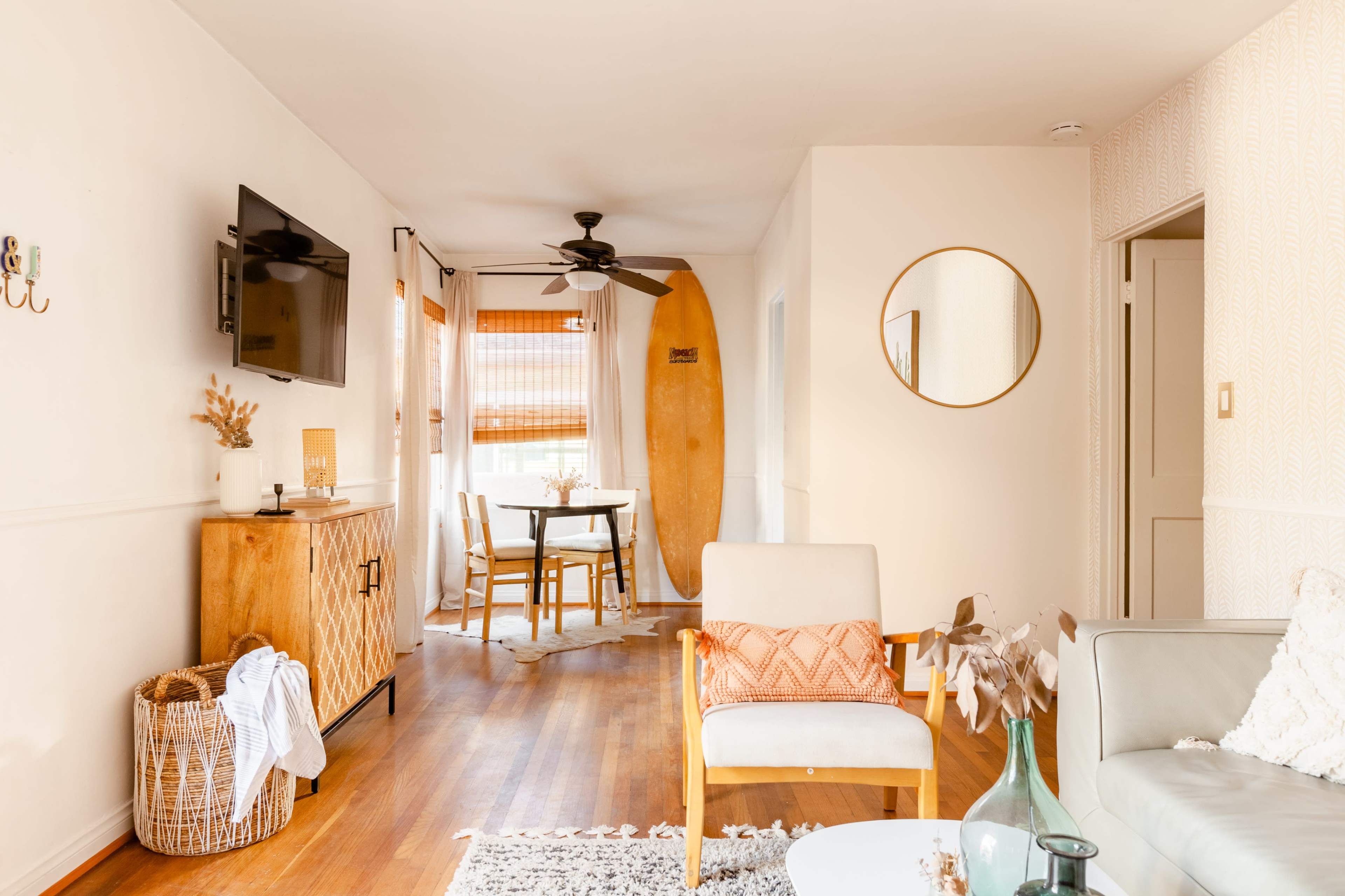 A bright living space with wooden floors, a surfboard mounted on the wall, and a dining area featuring a round table and four chairs.