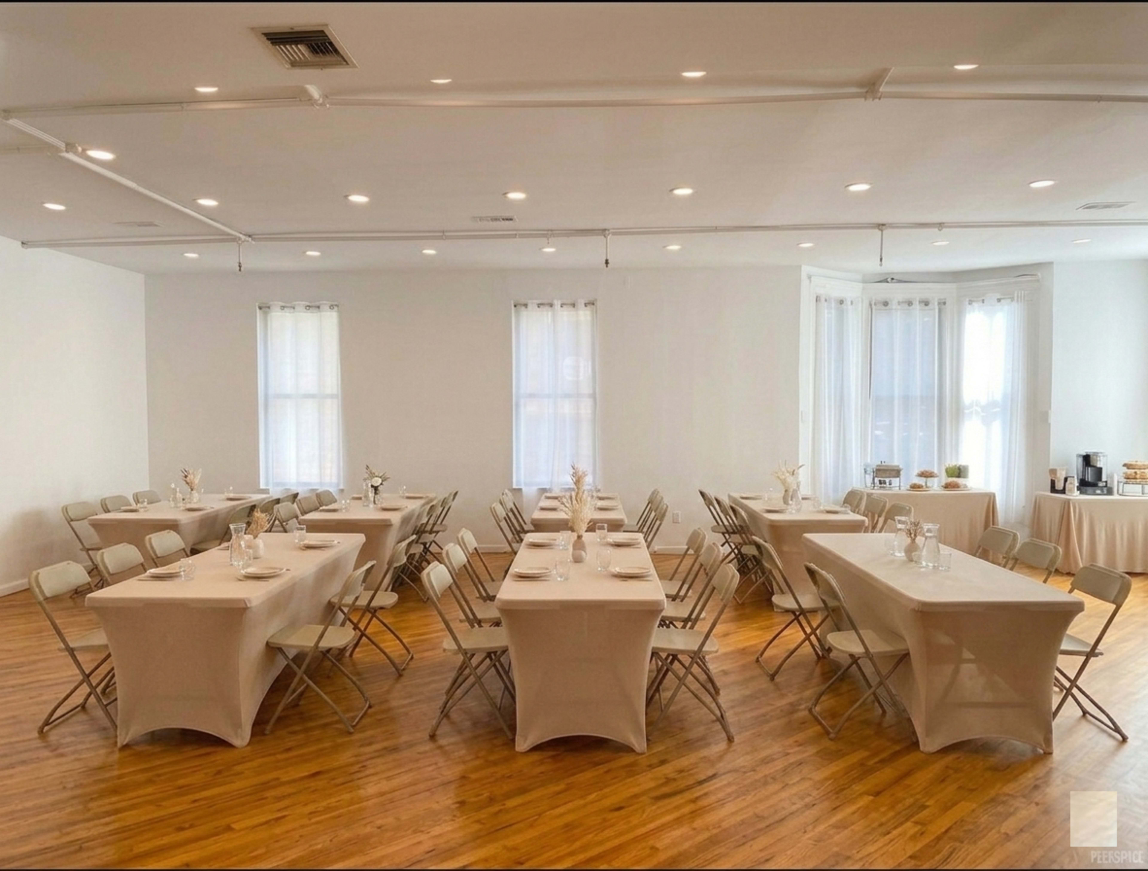 The image shows a spacious banquet hall set with several tables arranged in a U-shape, each covered with beige tablecloths and accompanied by folding chairs.