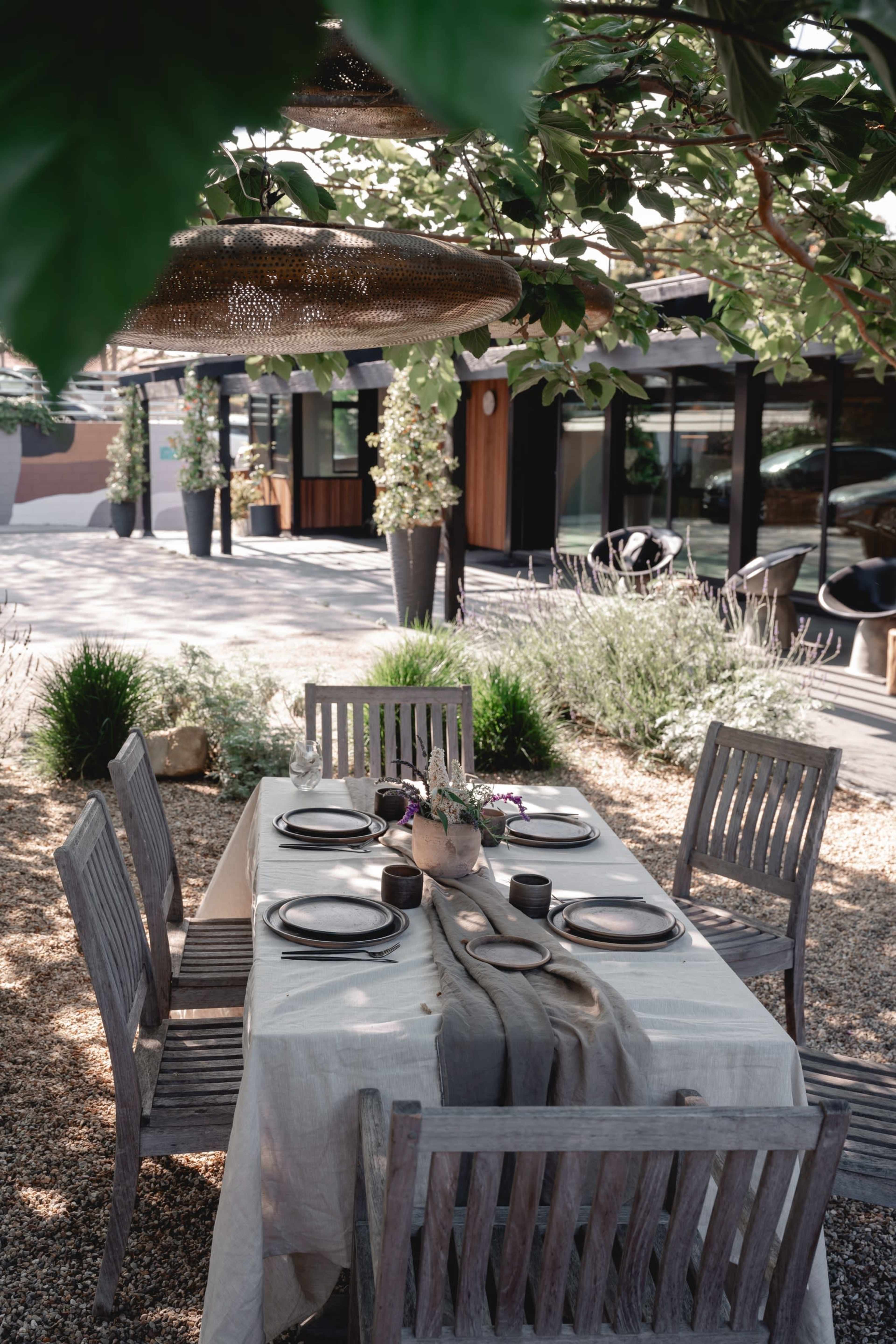 A comfortably set outdoor dining table with wooden chairs is surrounded by greenery and modern architecture in the background.
