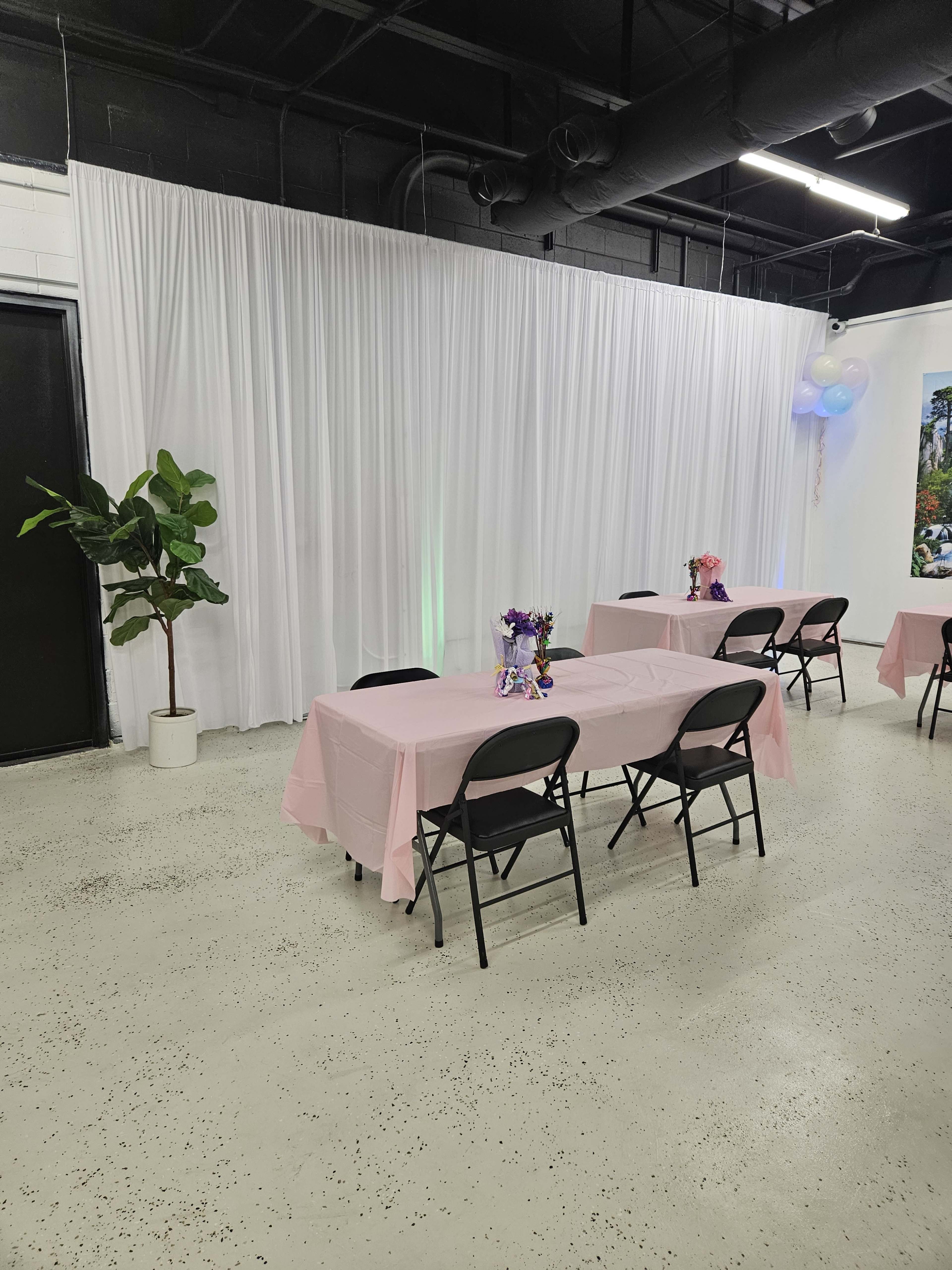 A room with pink-covered tables and a large white backdrop, featuring potted plants and decorative centerpieces.
