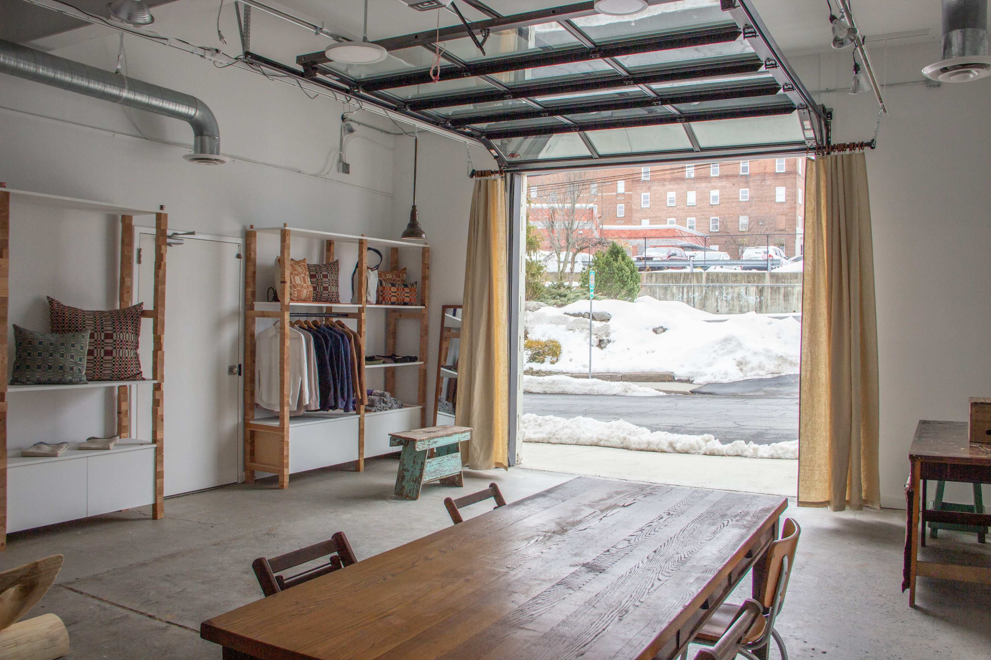 The image shows a spacious interior of a modern garage or studio with a large open door, a wooden table, shelves of clothing, and snow outside.