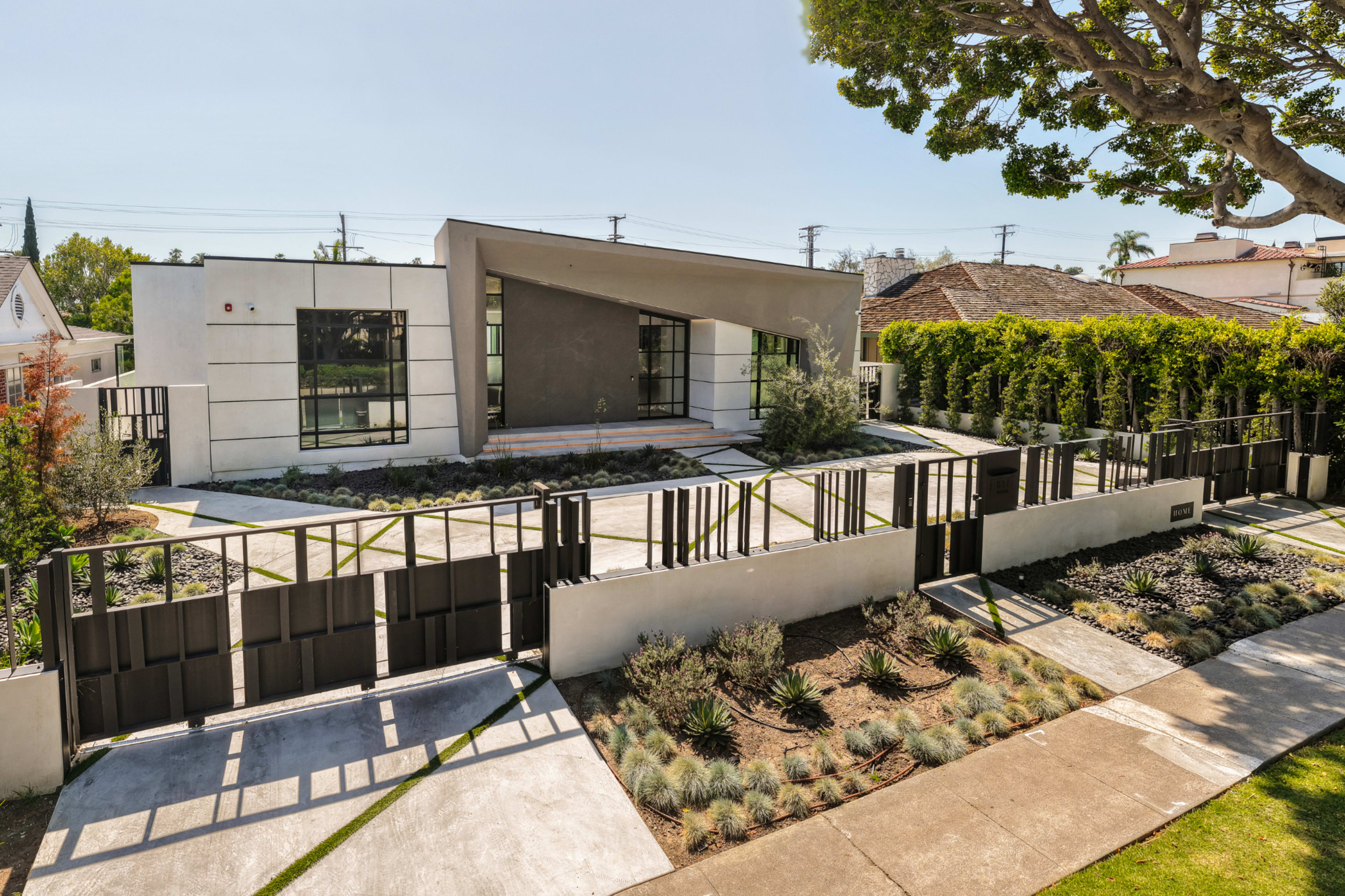 The image shows a modern house with a distinctive architectural design, featuring large windows, a landscaped front yard, and a gated entrance.
