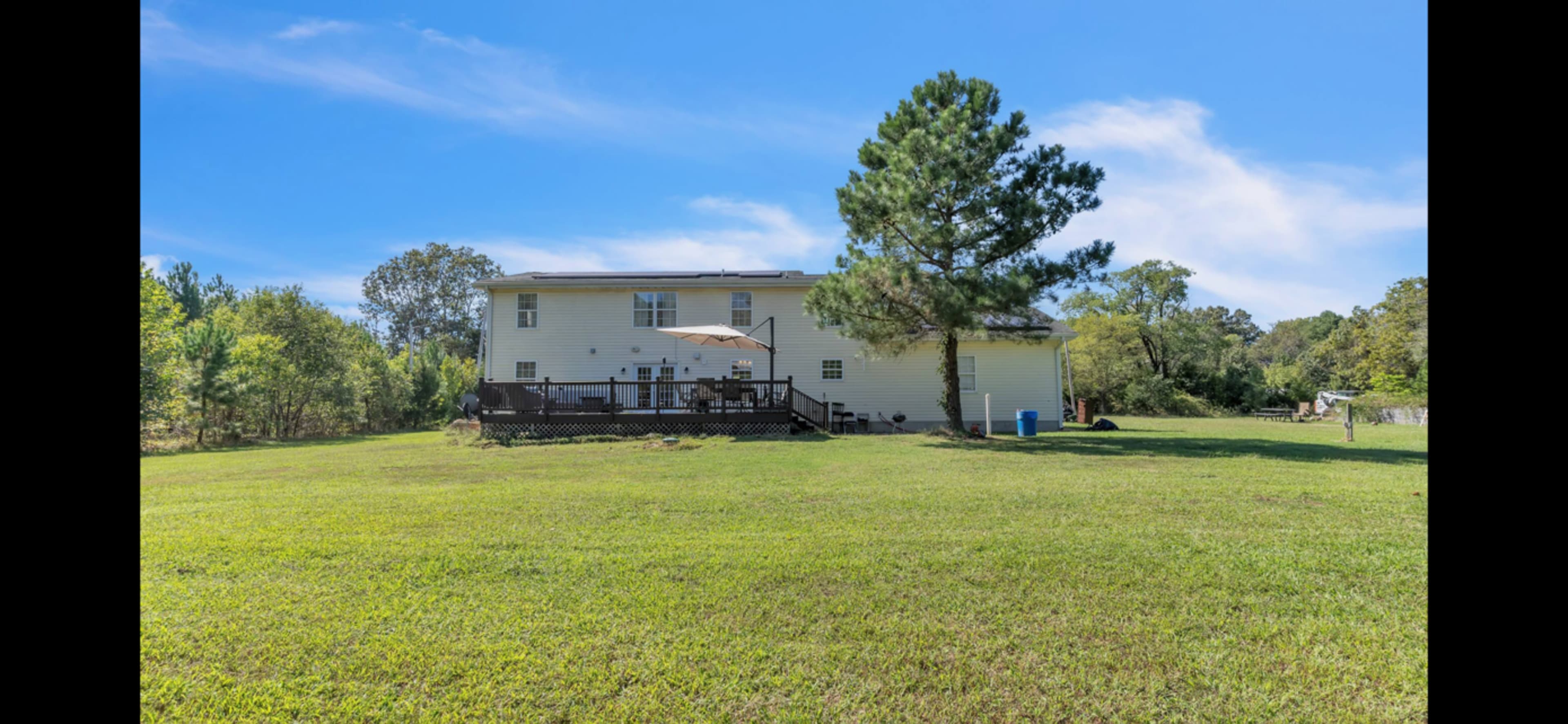 A two-story house with a deck and an umbrella is situated on a grassy yard surrounded by trees.