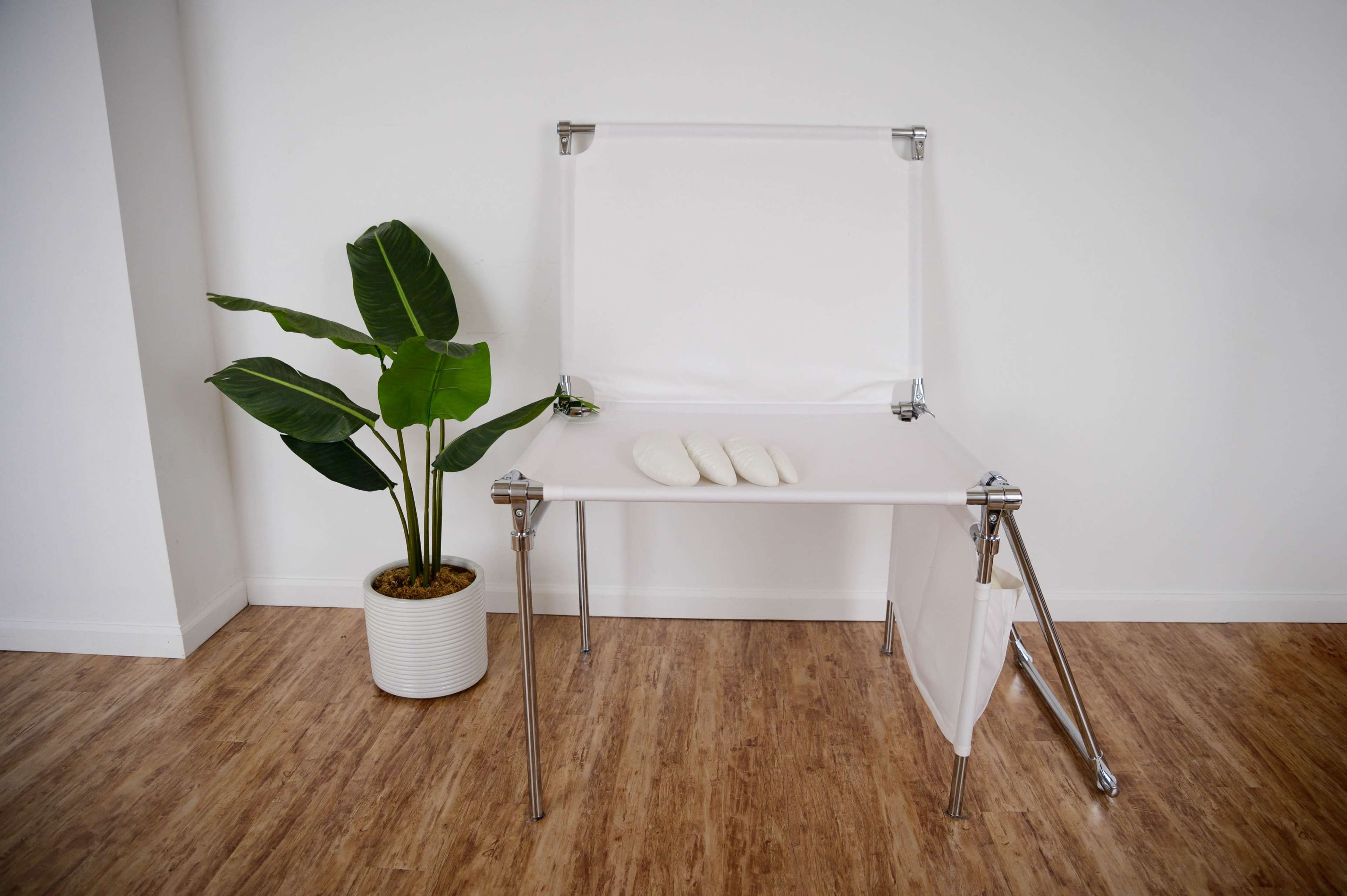 A simple white table with three pillows rests against a plain wall, alongside a large green plant in a white pot.
