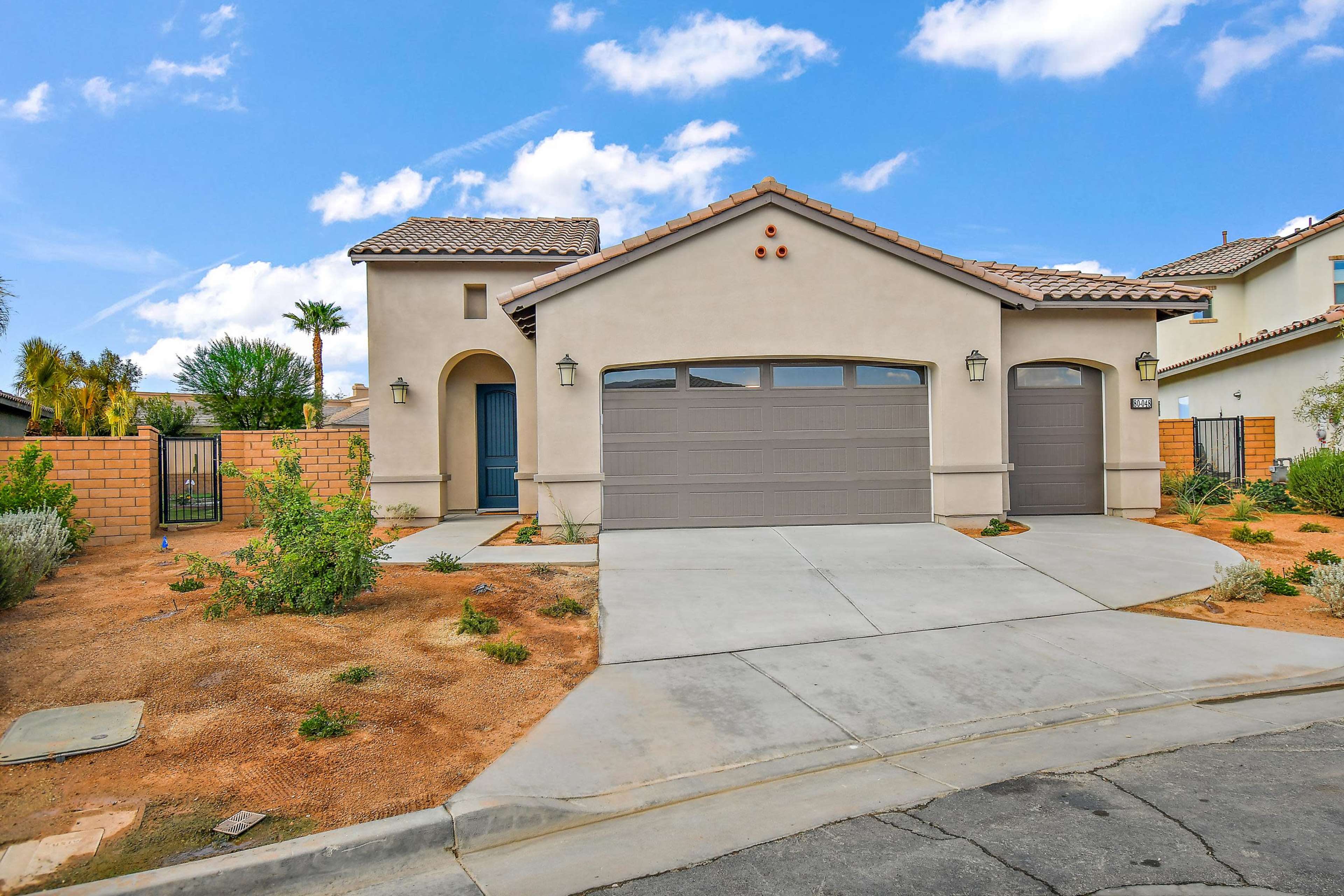 A single-story house with a beige exterior and a dual garage sits on a corner lot, surrounded by sparse landscaping and a driveway.