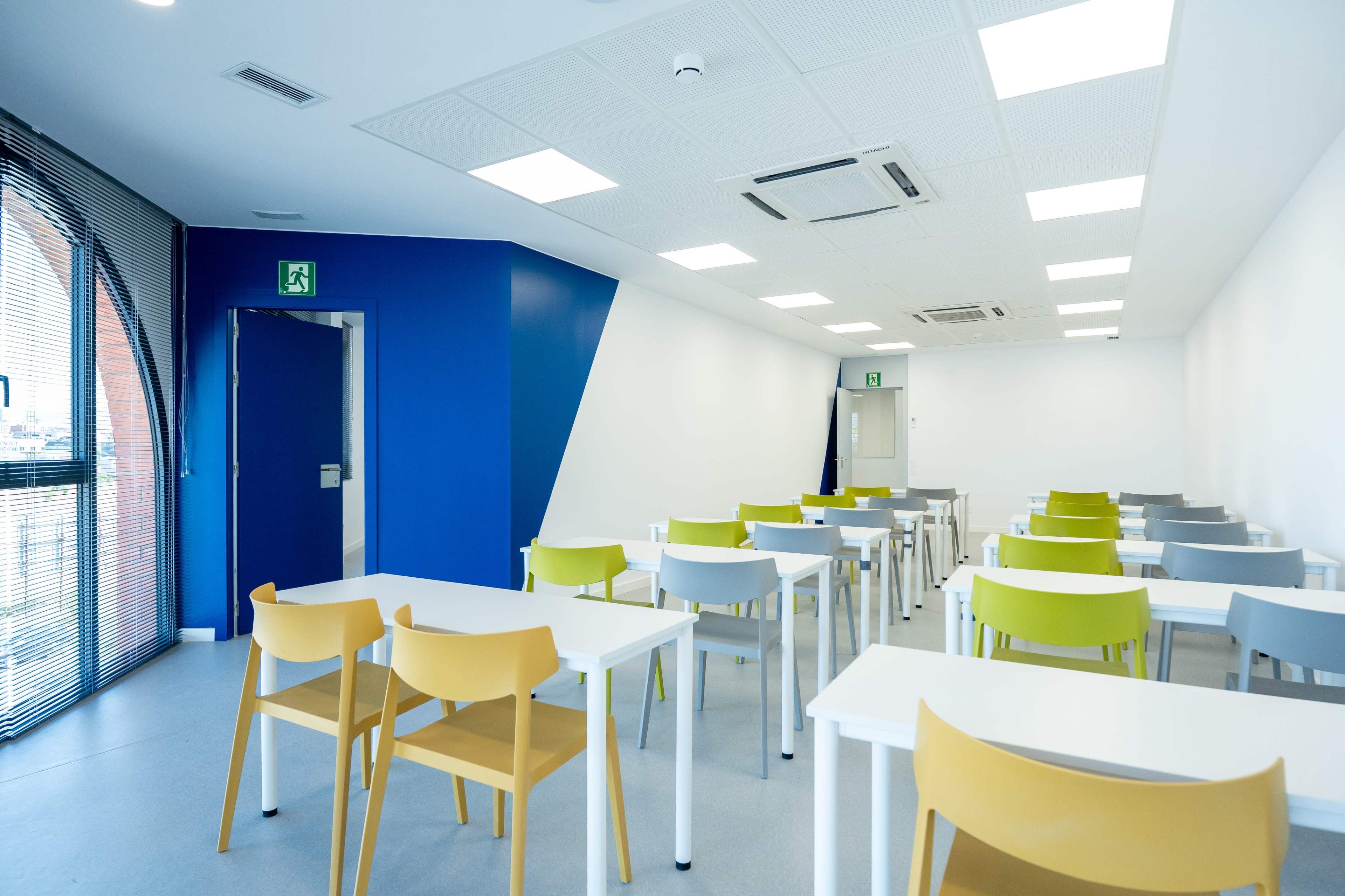 The image shows a modern classroom with rows of tables and chairs in various colors, featuring large windows and bright lighting.