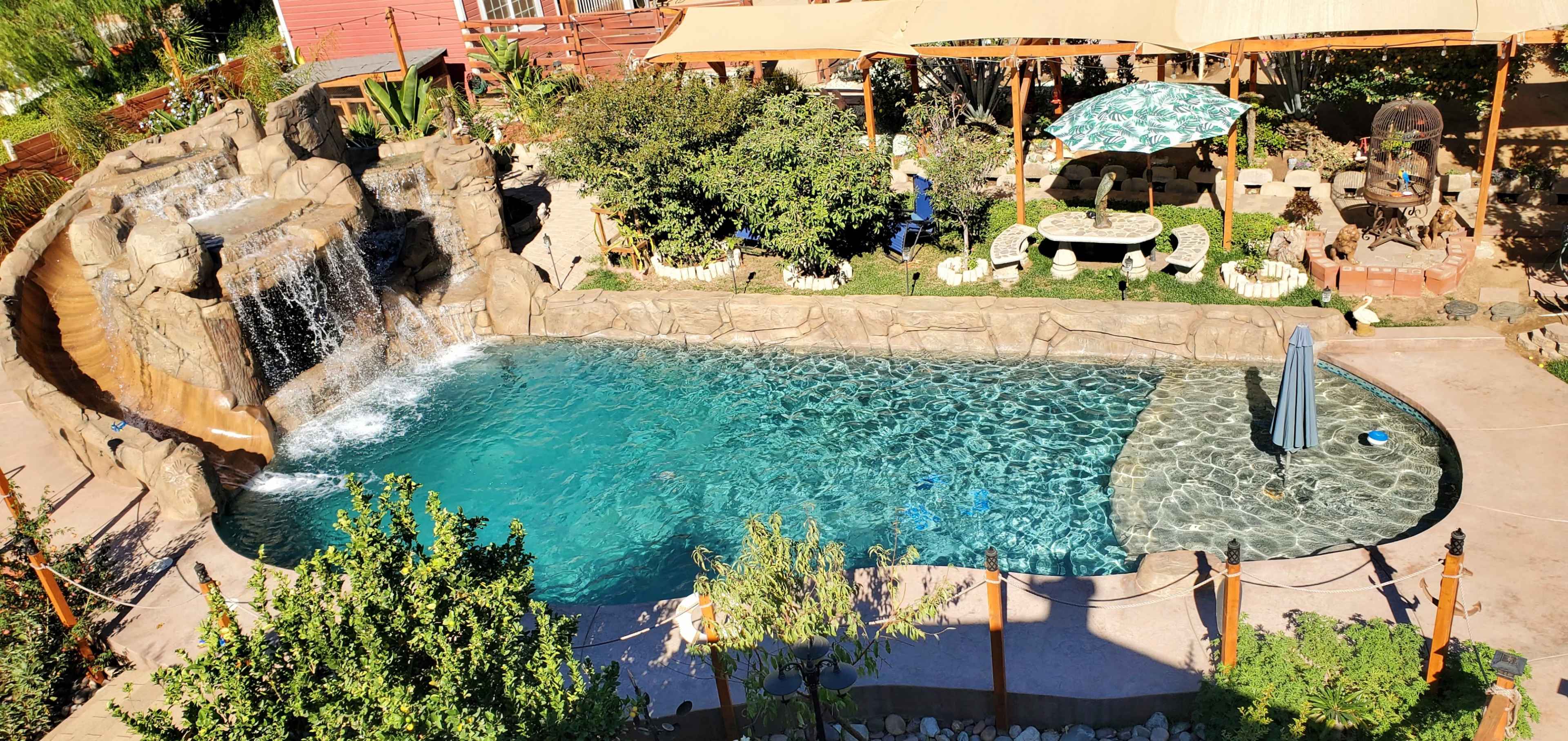 The image shows a backyard pool with a rock waterfall, surrounded by lush greenery and seating areas.