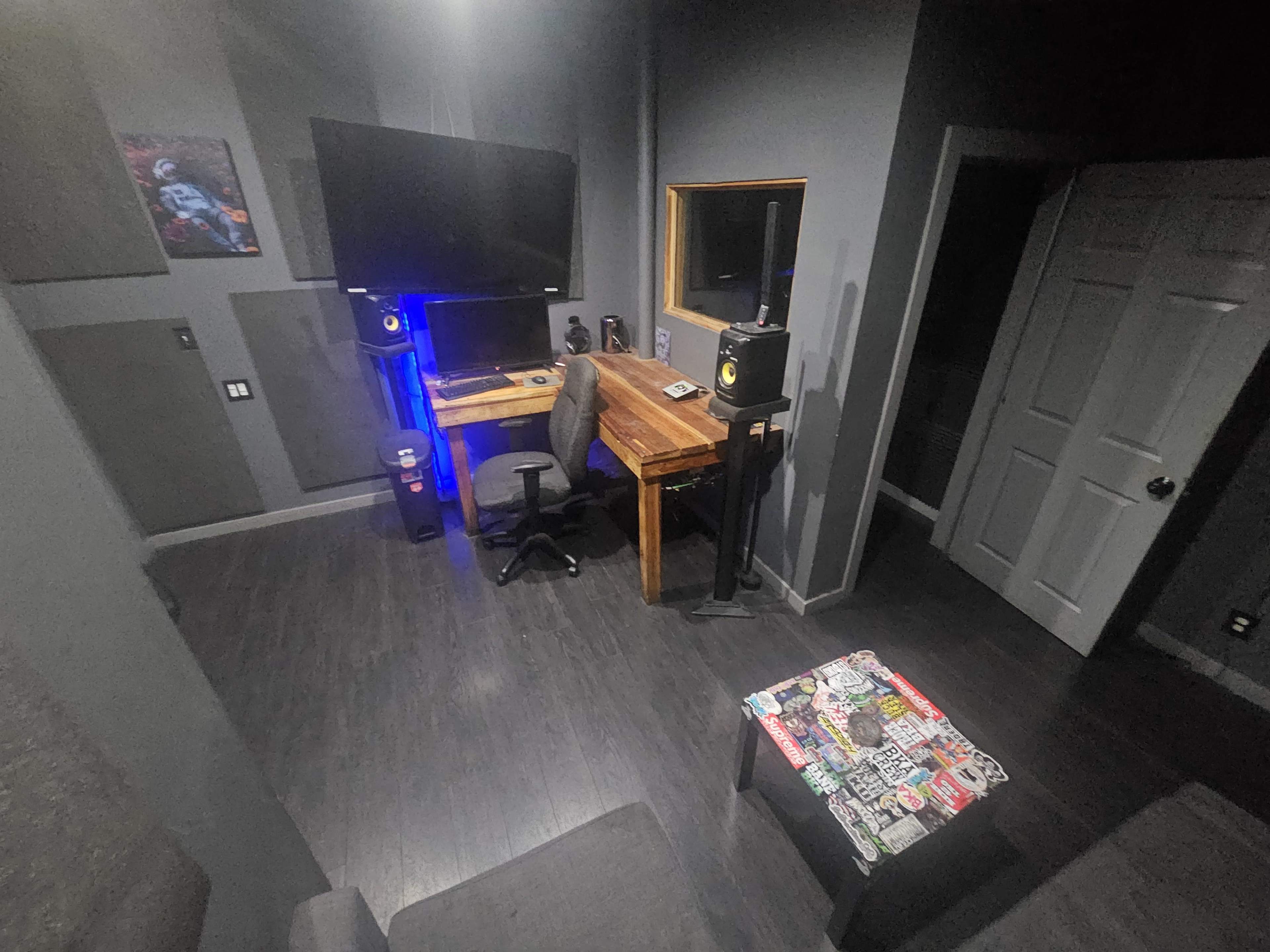 The image shows a dimly lit recording studio featuring a desk with a computer, speakers, and a chair, along with a couch and a wall-mounted television.