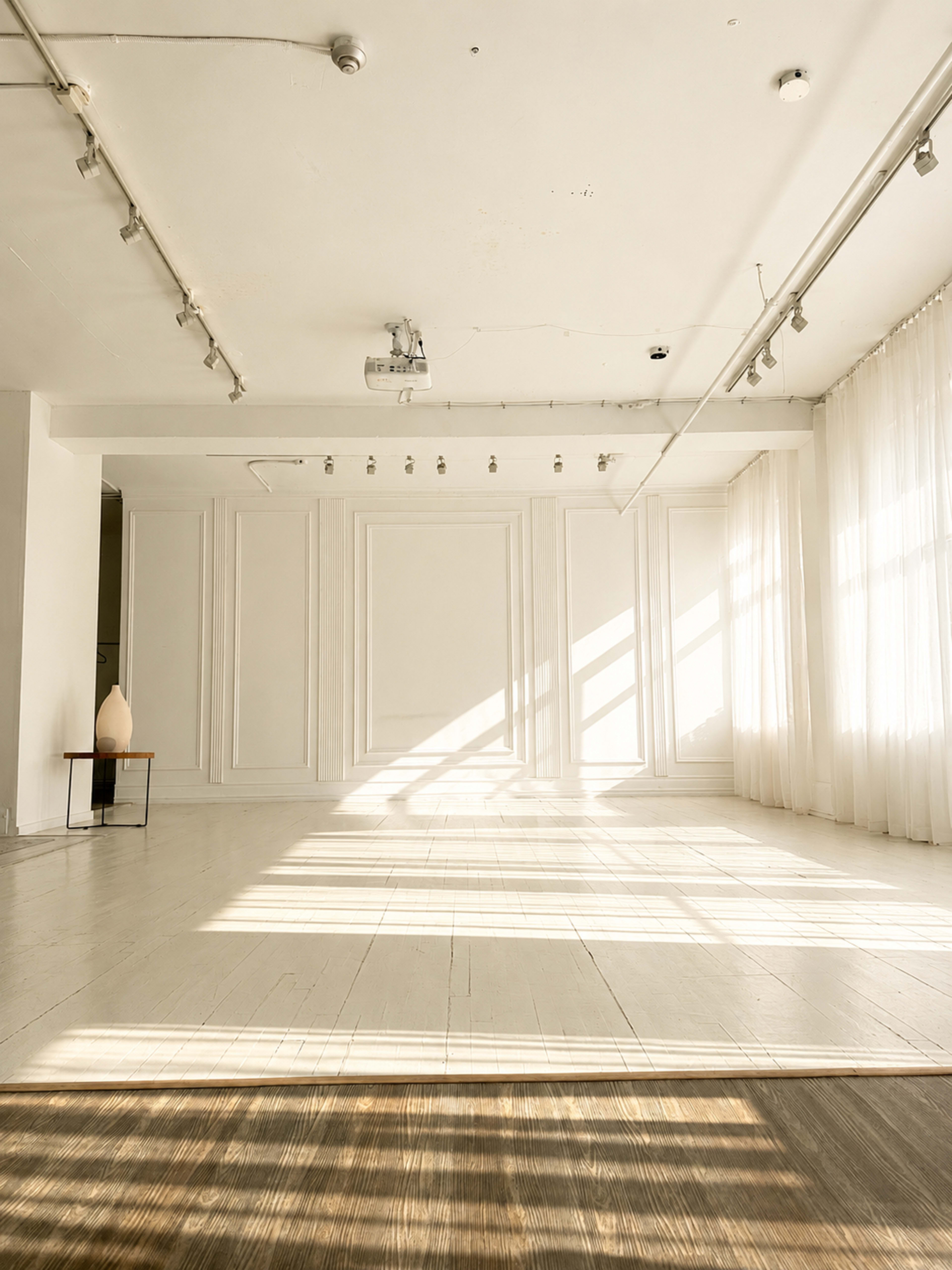 The image shows a sunlit, empty room featuring white walls, wooden flooring, and large windows with sheer curtains.