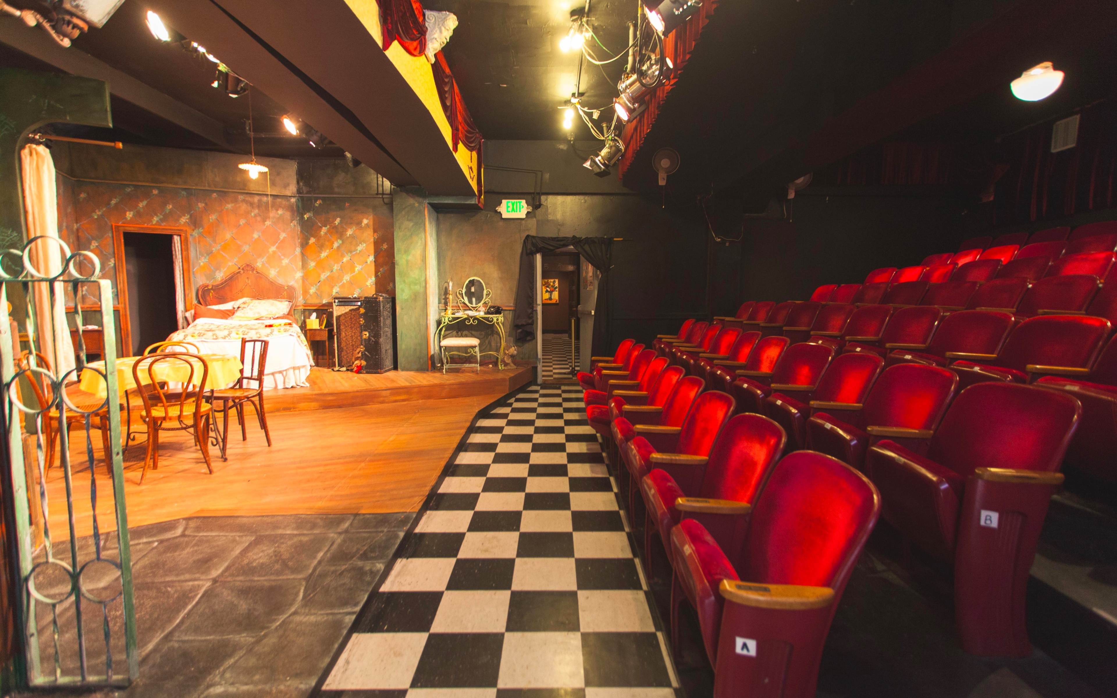 A small theater with red seats facing a stage that features a bed and vintage furniture in a dimly lit space.