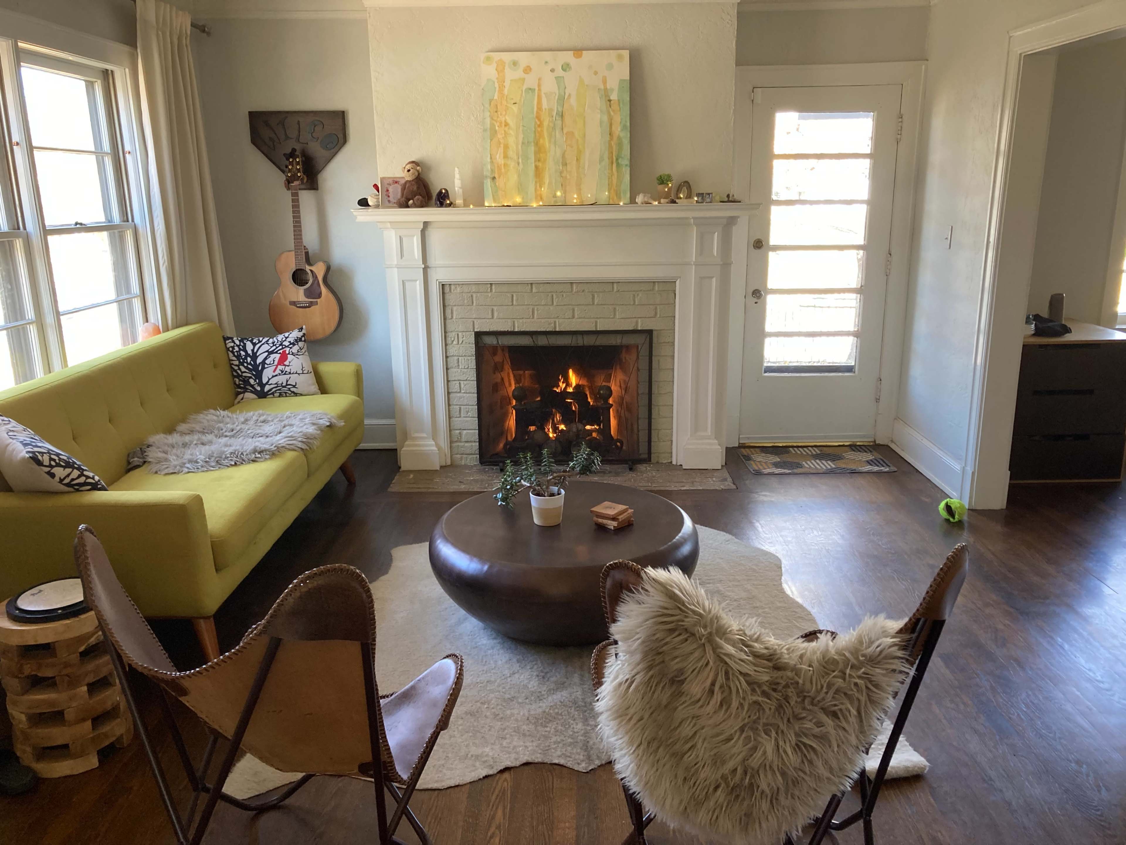 The living room features a yellow sofa, two leather chairs with fur throws, a round coffee table, and a fireplace with a fire burning in it.