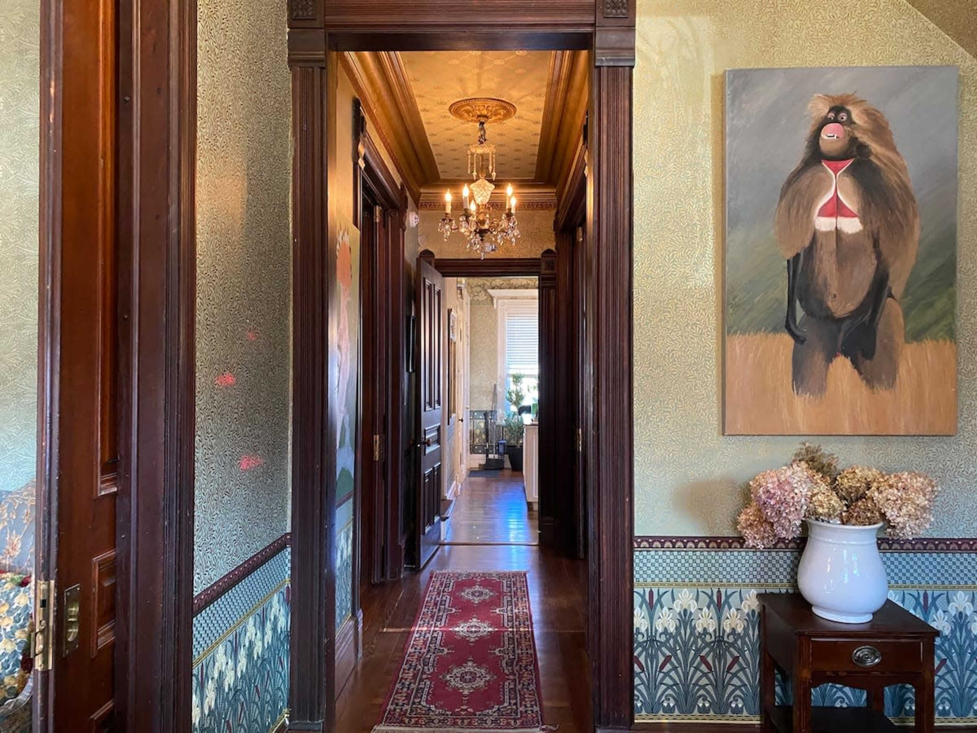 A hallway with ornate wallpaper leads to a room with a chandelier, featuring a large painting of a baboon on the wall.