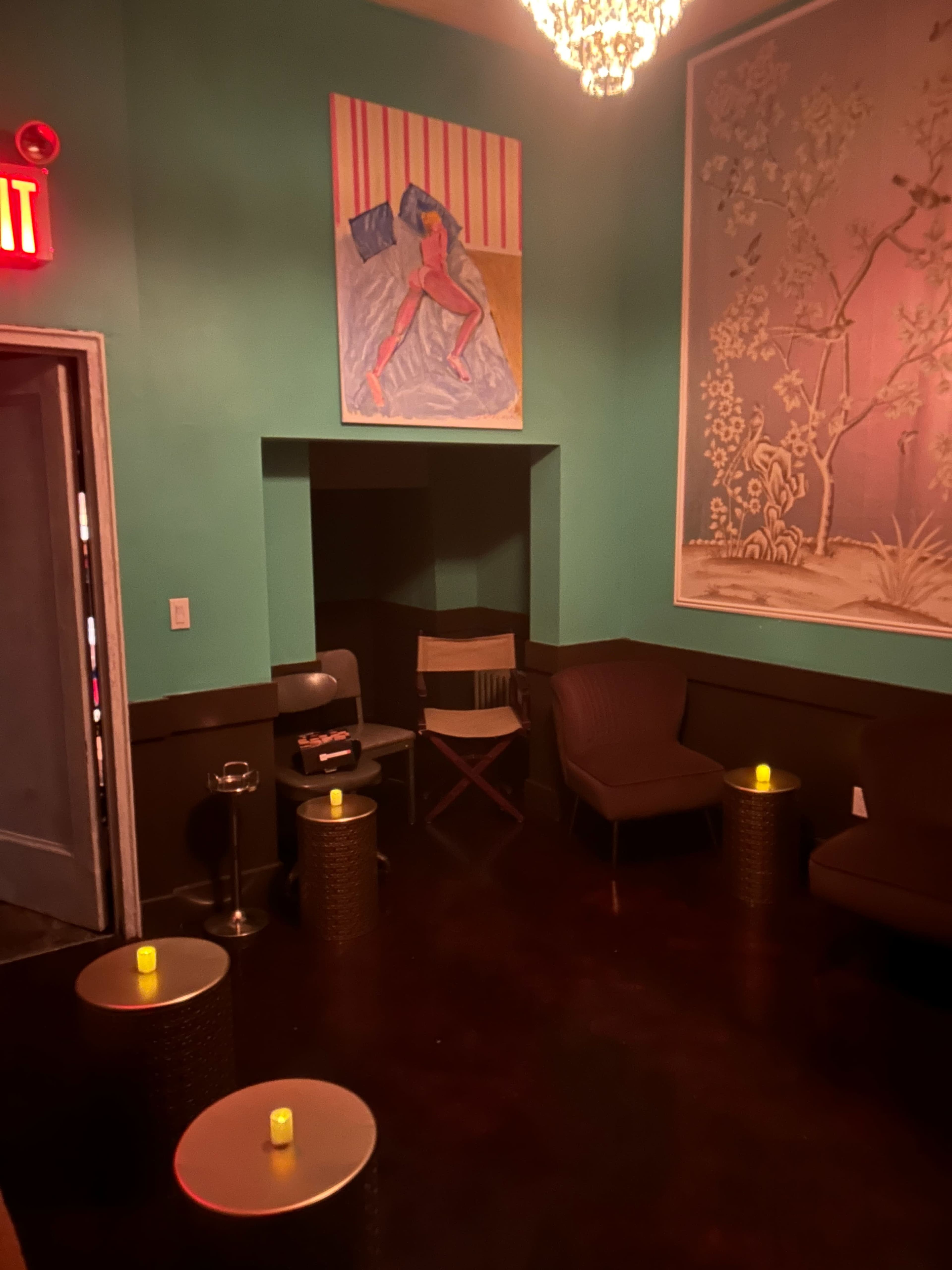 The image shows a dimly lit room with turquoise walls, a large artwork, and several small tables with candles, along with a few chairs arranged in the space.