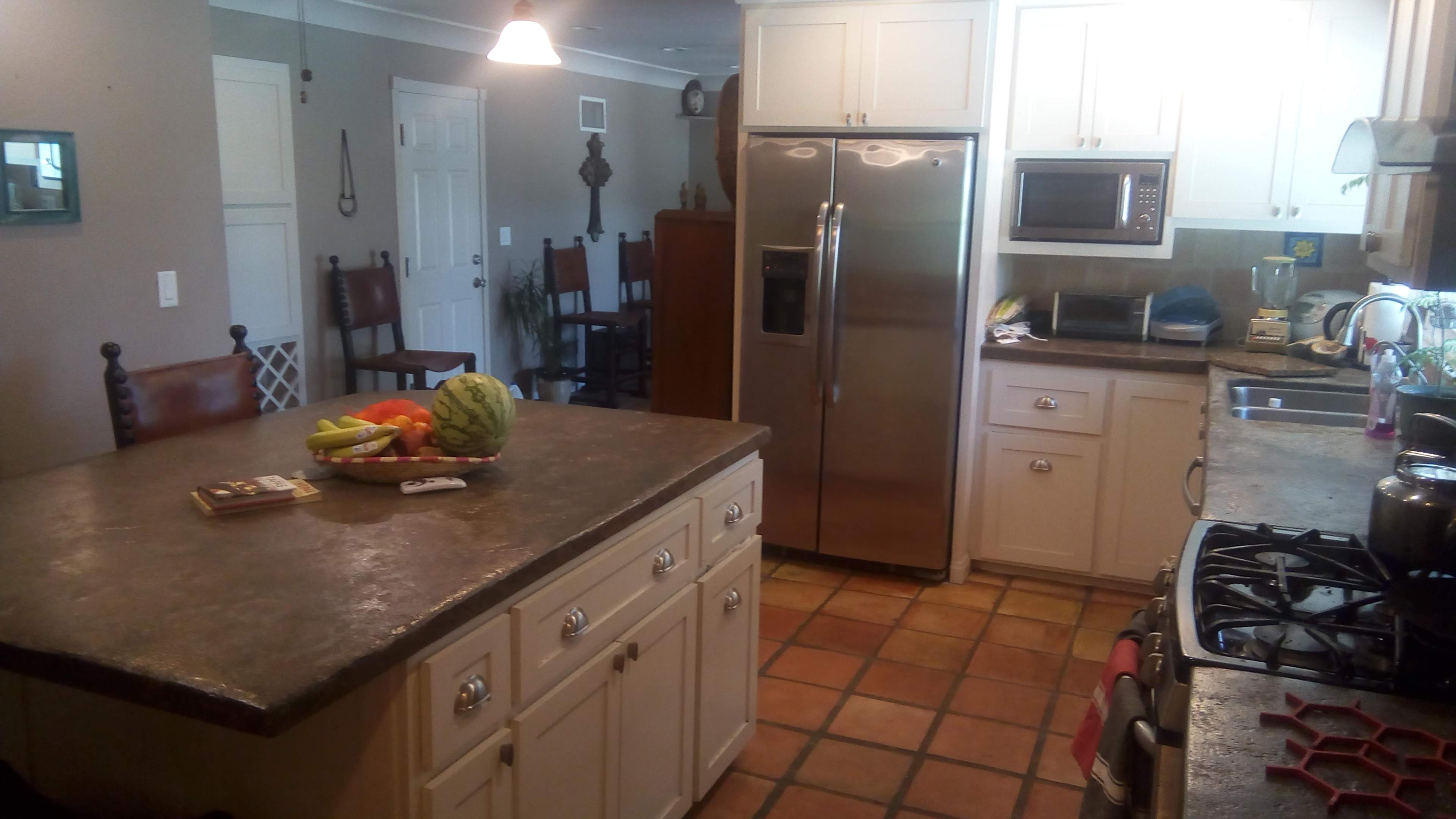 A kitchen with a central island topped with a bowl of fruit, stainless steel appliances, and tiled floors.