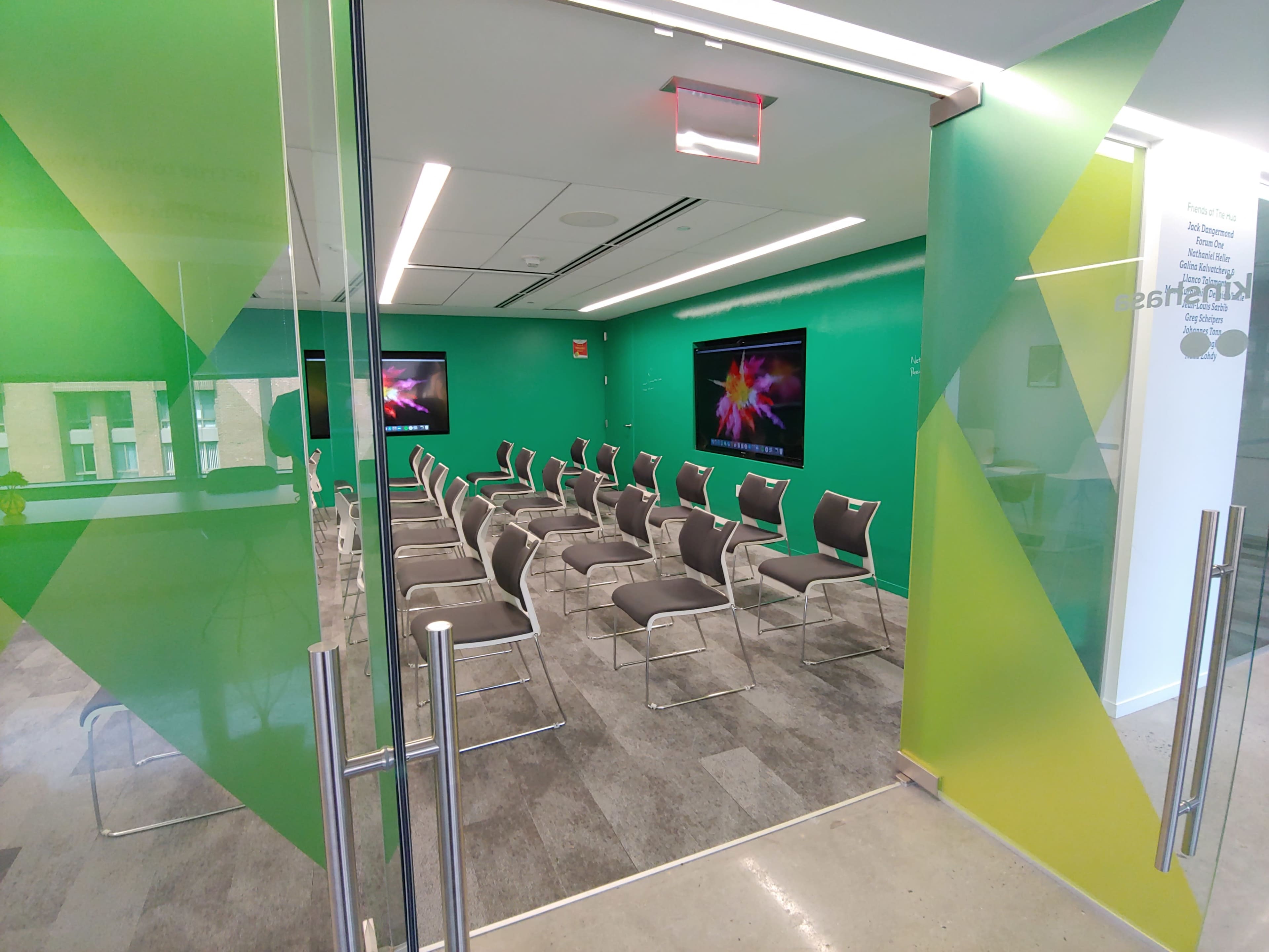 A modern conference room with green walls, two screens, and several rows of gray chairs arranged for a presentation.
