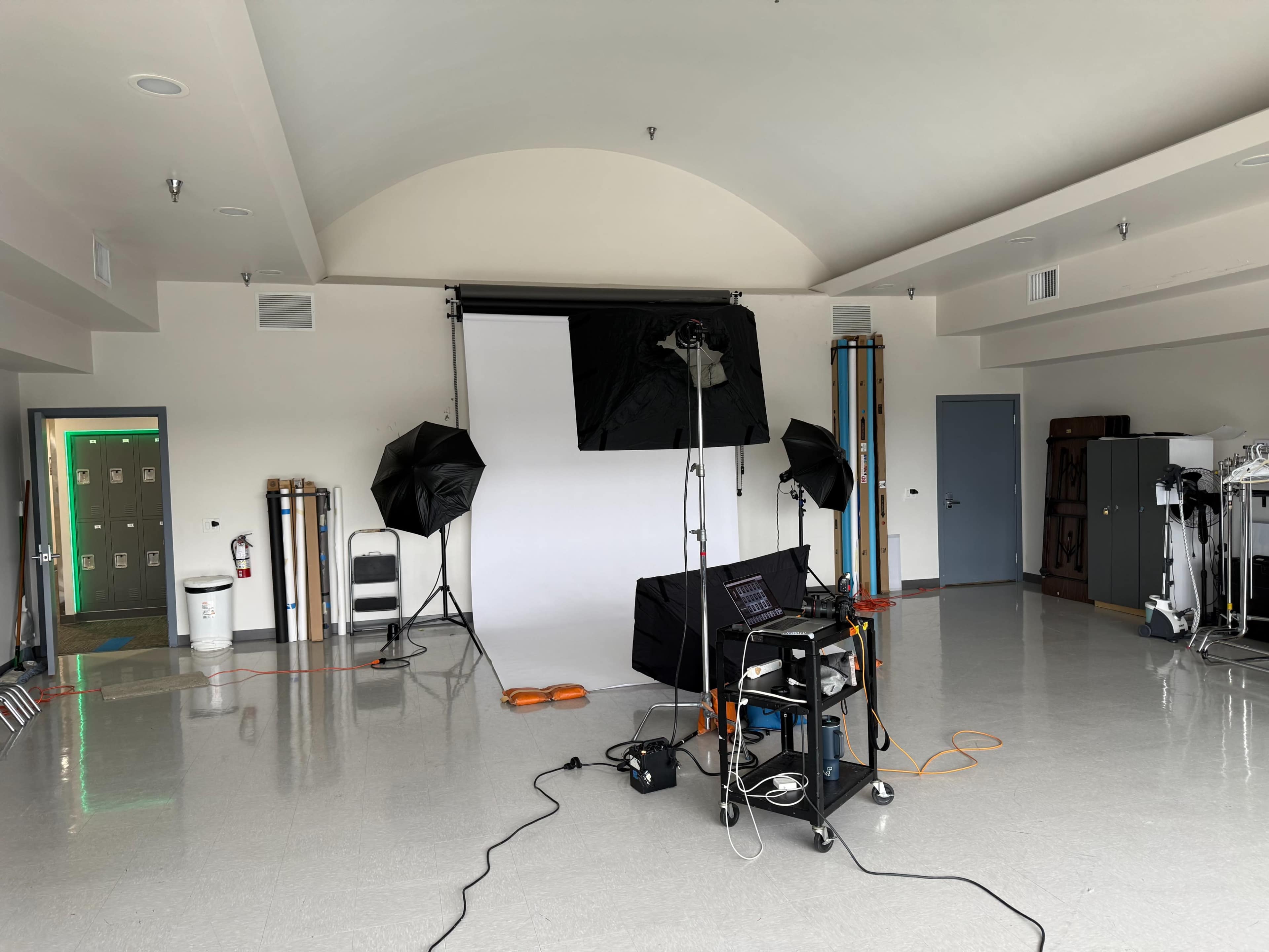 The image shows a photography studio setup with softbox lights, a backdrop, and a laptop on a portable table, all positioned in a spacious, empty room.